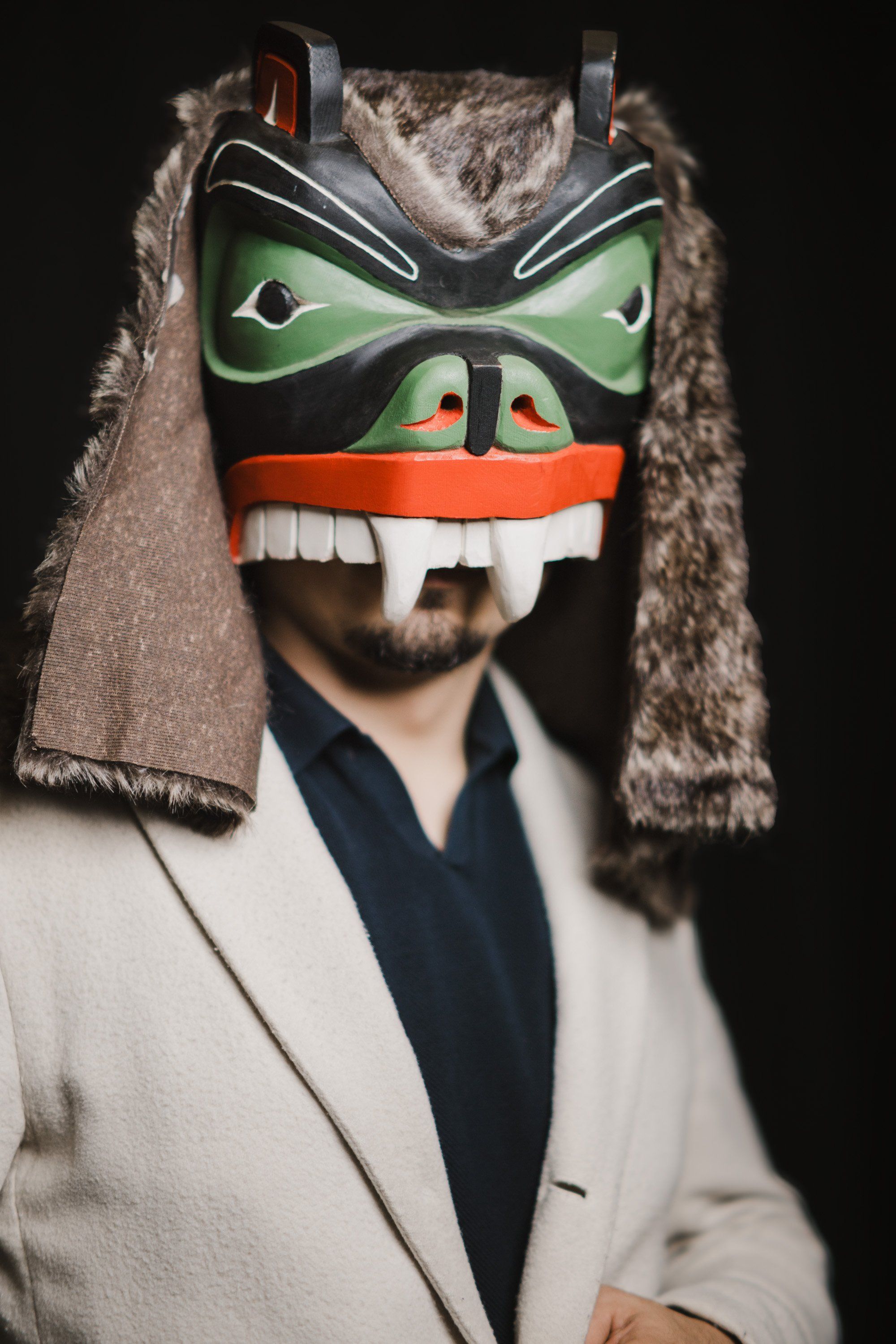 Person in cream blazer wearing carved wooden mask with green eye panels, orange mouth, and fur-trimmed regalia against black background