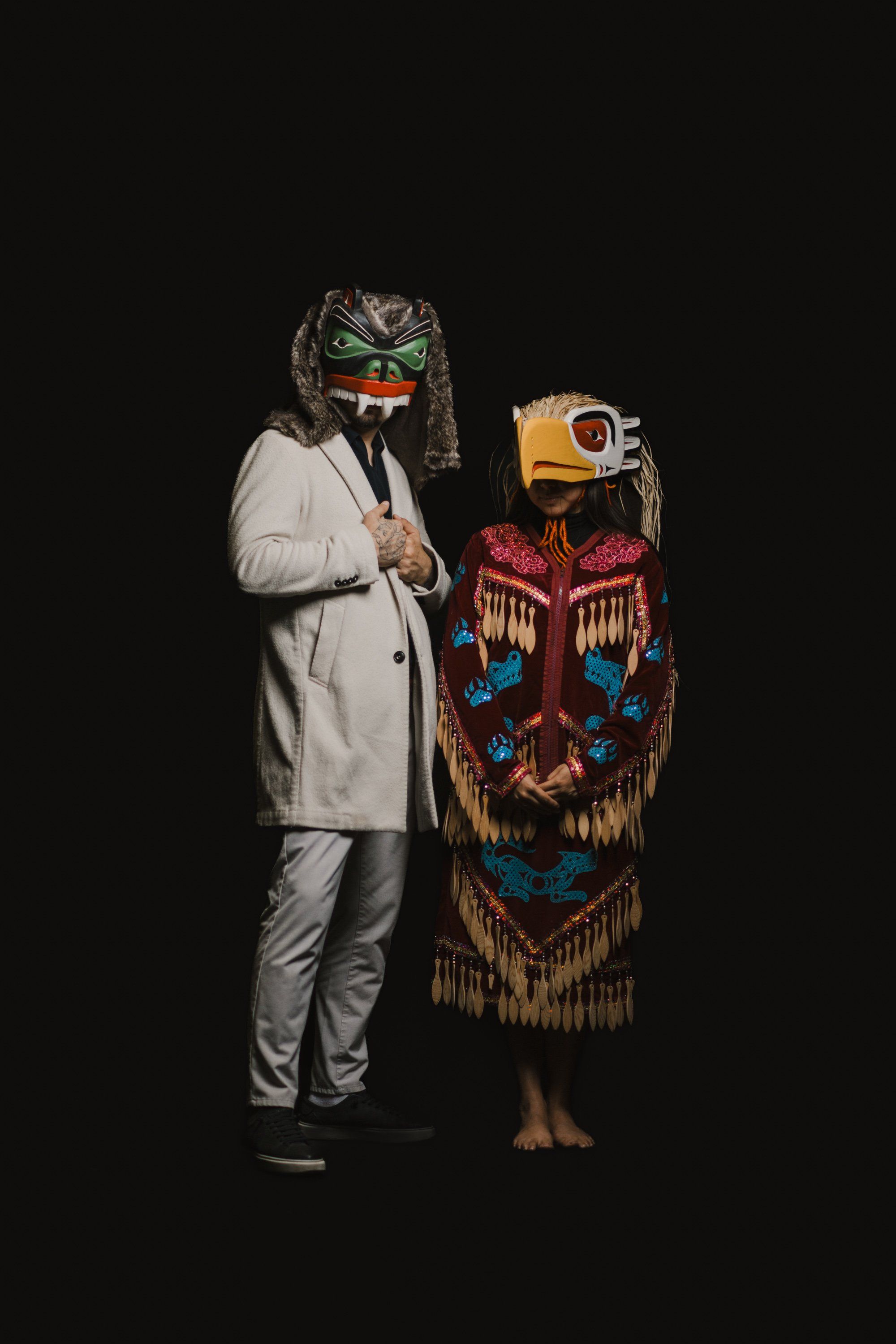 Person in light grey suit and carved mask stands beside figure in red and black regalia with traditional Northwest Coast design against black background