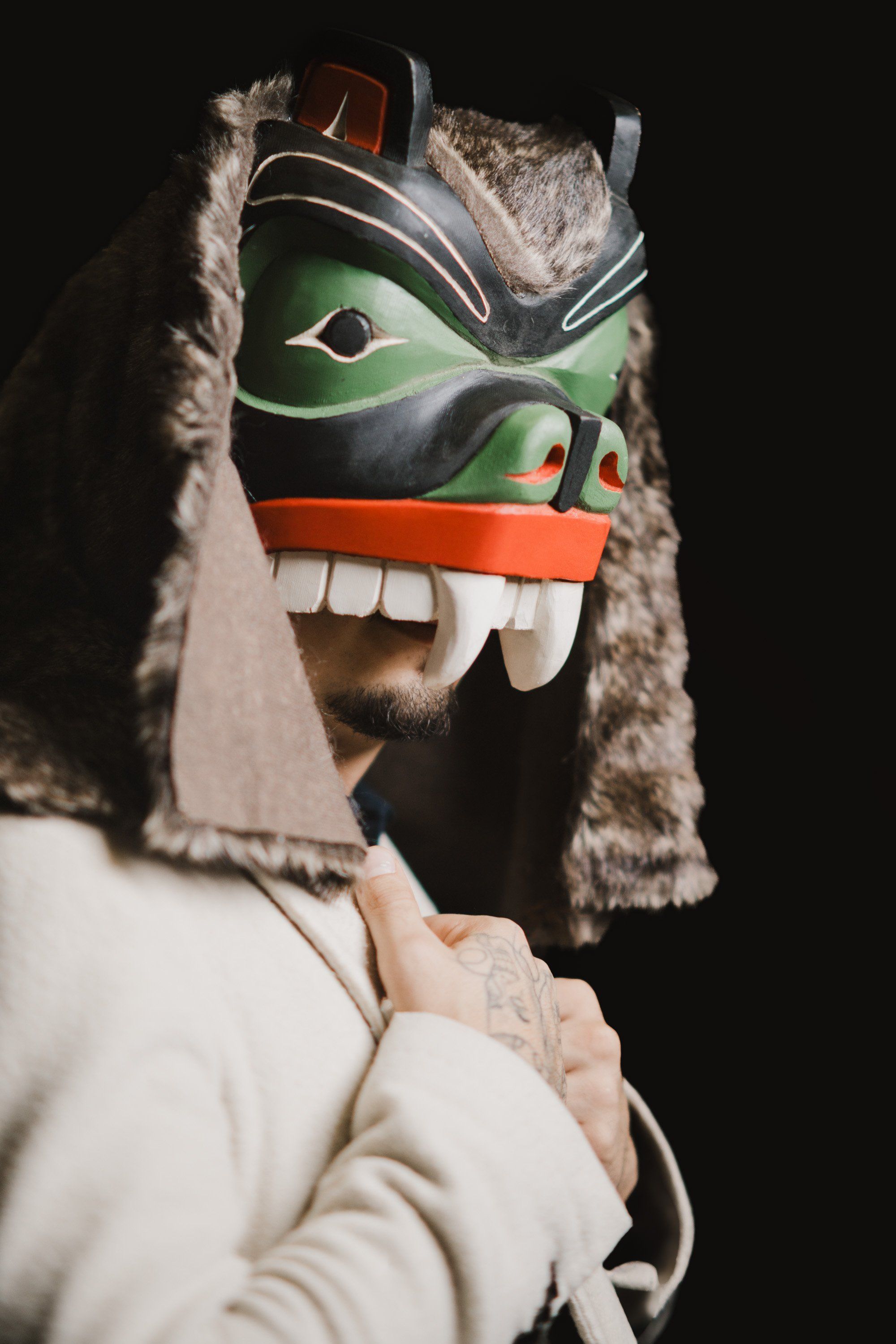 Person wearing traditional Coast Salish wolf mask with green face paint, fur collar, and white teeth against black background