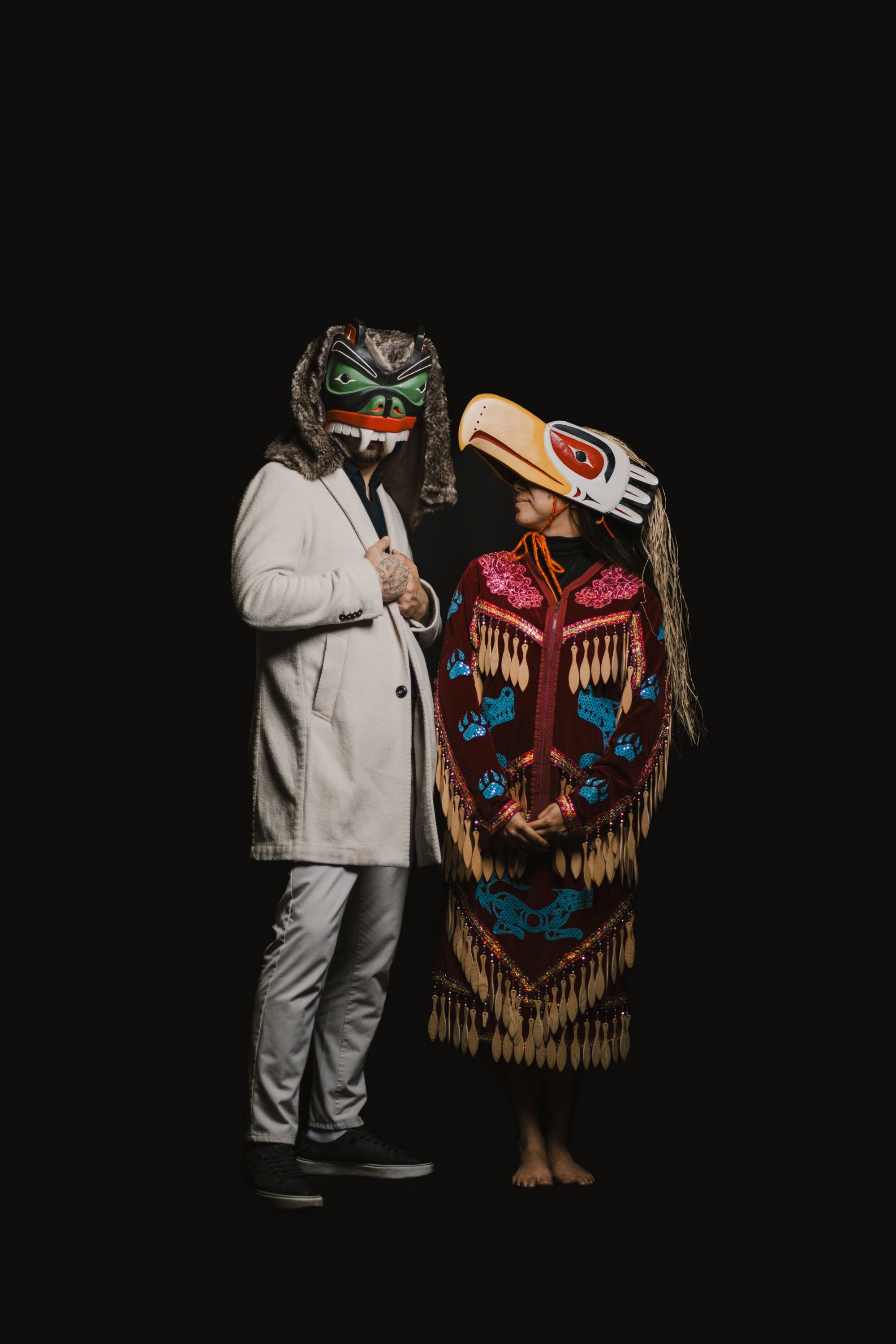 Person in light blazer and mask stands beside figure in ornate red and blue regalia holding traditional carved wooden mask, both posed against black background