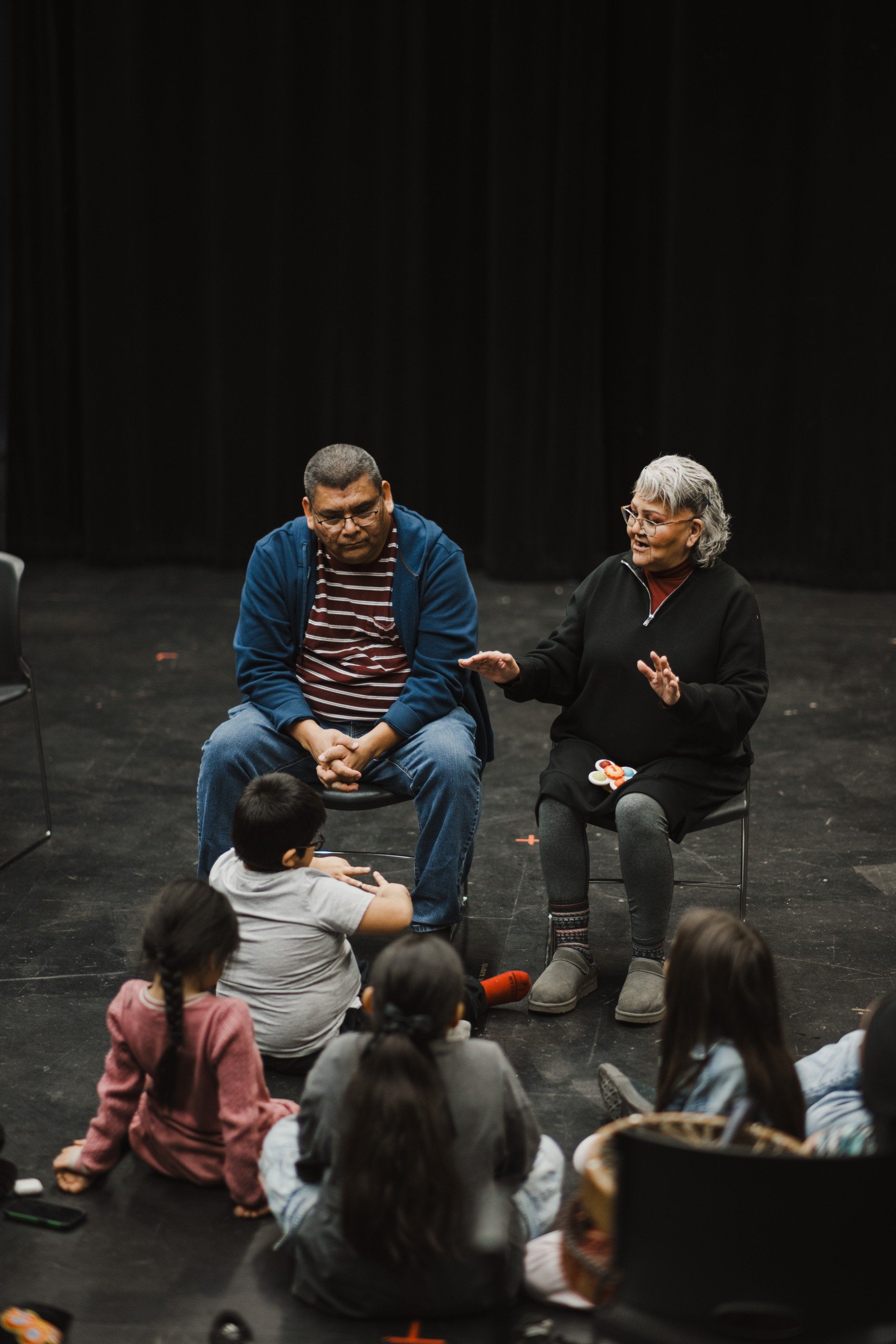 An elder and man in striped shirt sit facing children seated on a dark stage, engaged in intimate conversation during a Coast Salish cultural celebration