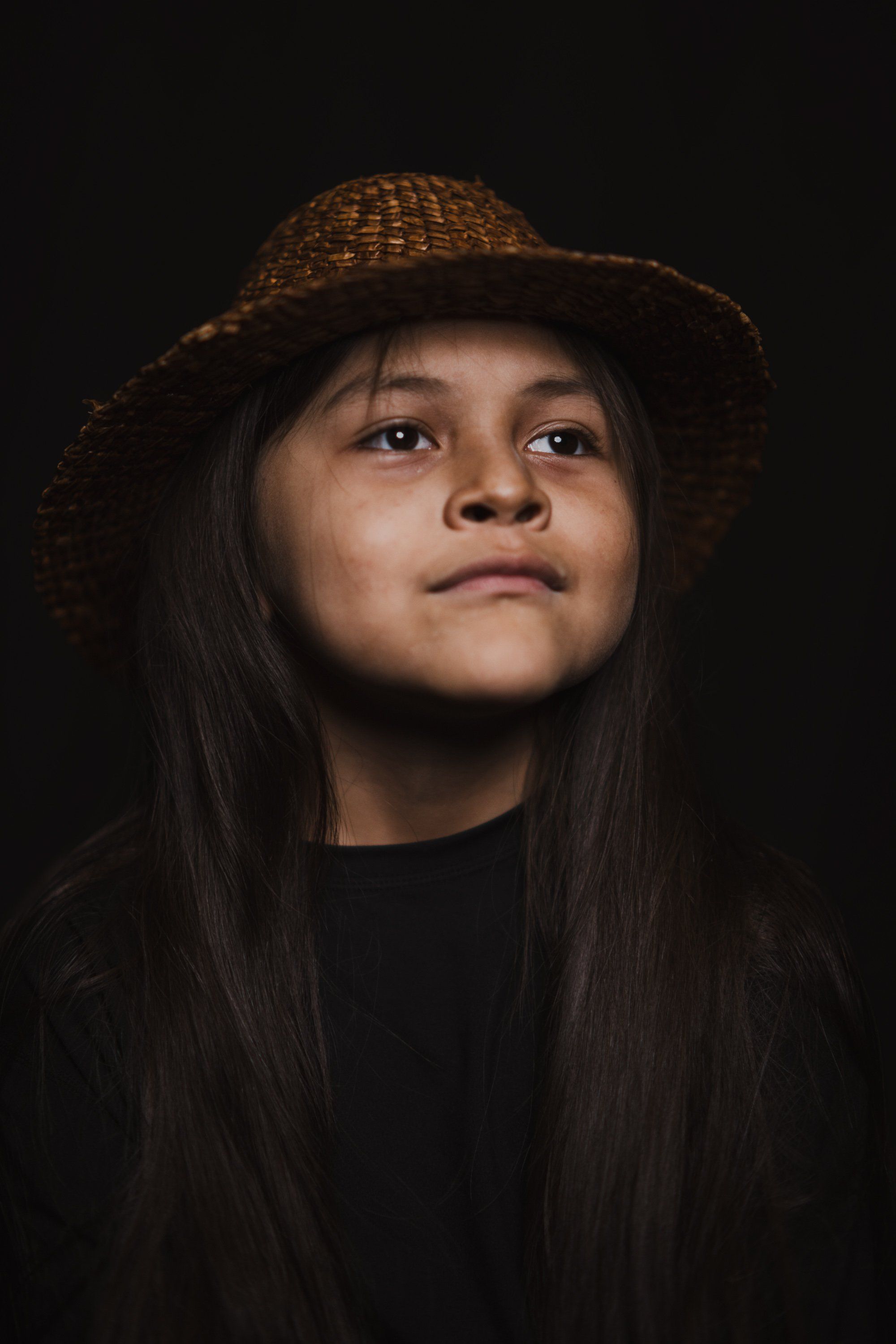 Young person wearing a woven hat and black clothing, gazing upward against a dark background with warm, directional lighting