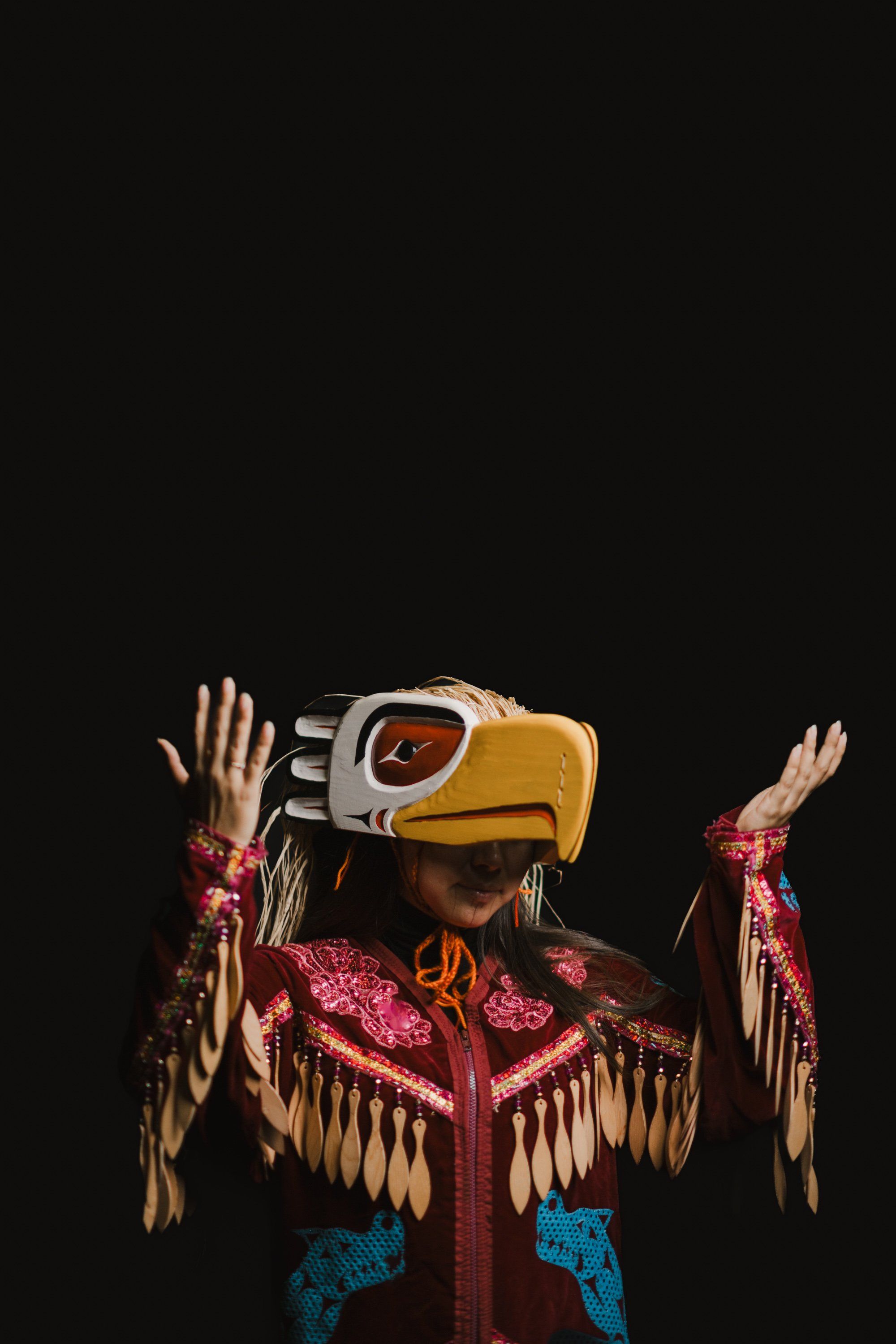Coast Salish dancer in golden eagle regalia with red button blanket and carved mask, arms raised against black background