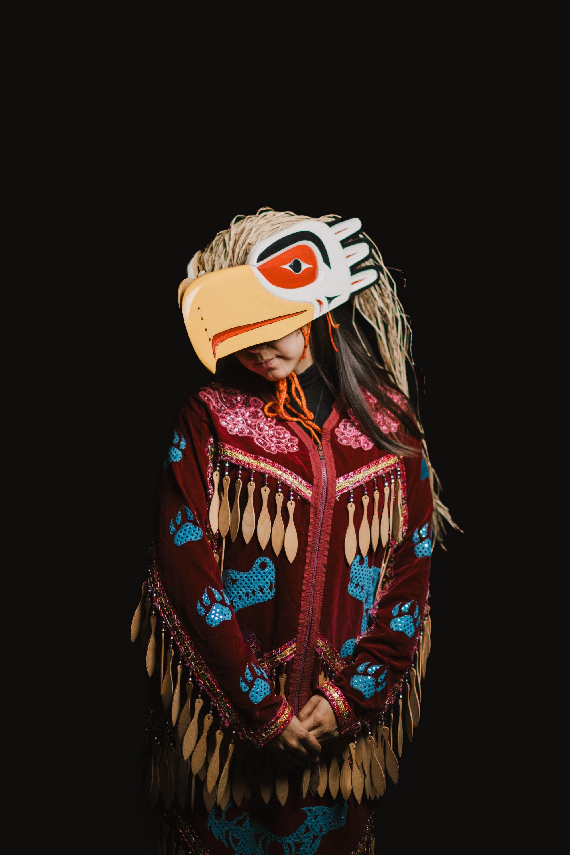 Young dancer in traditional Coast Salish regalia with eagle headdress and burgundy button blanket against black background