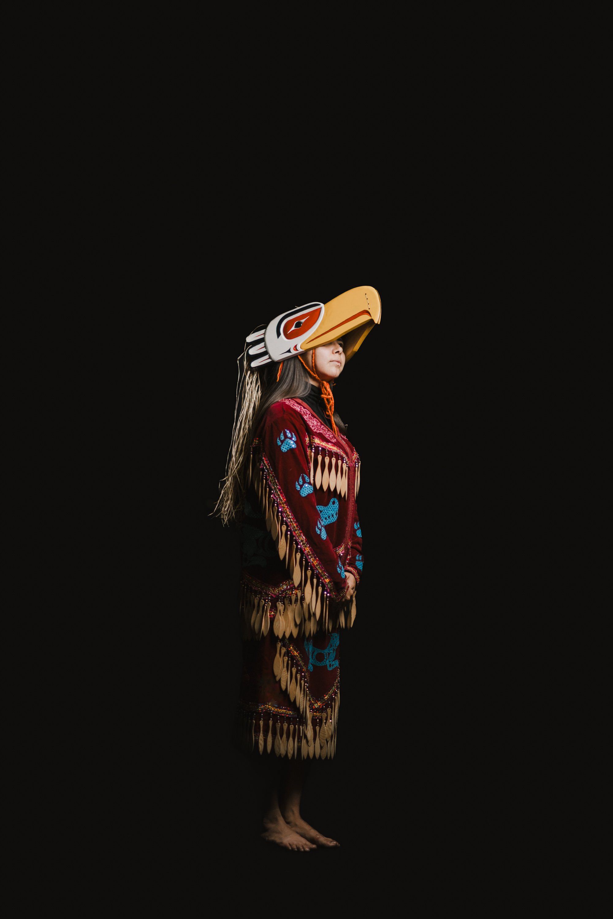 Coast Salish dancer in traditional regalia with carved wooden mask and red blanket stands against black background, arms raised in ceremonial pose