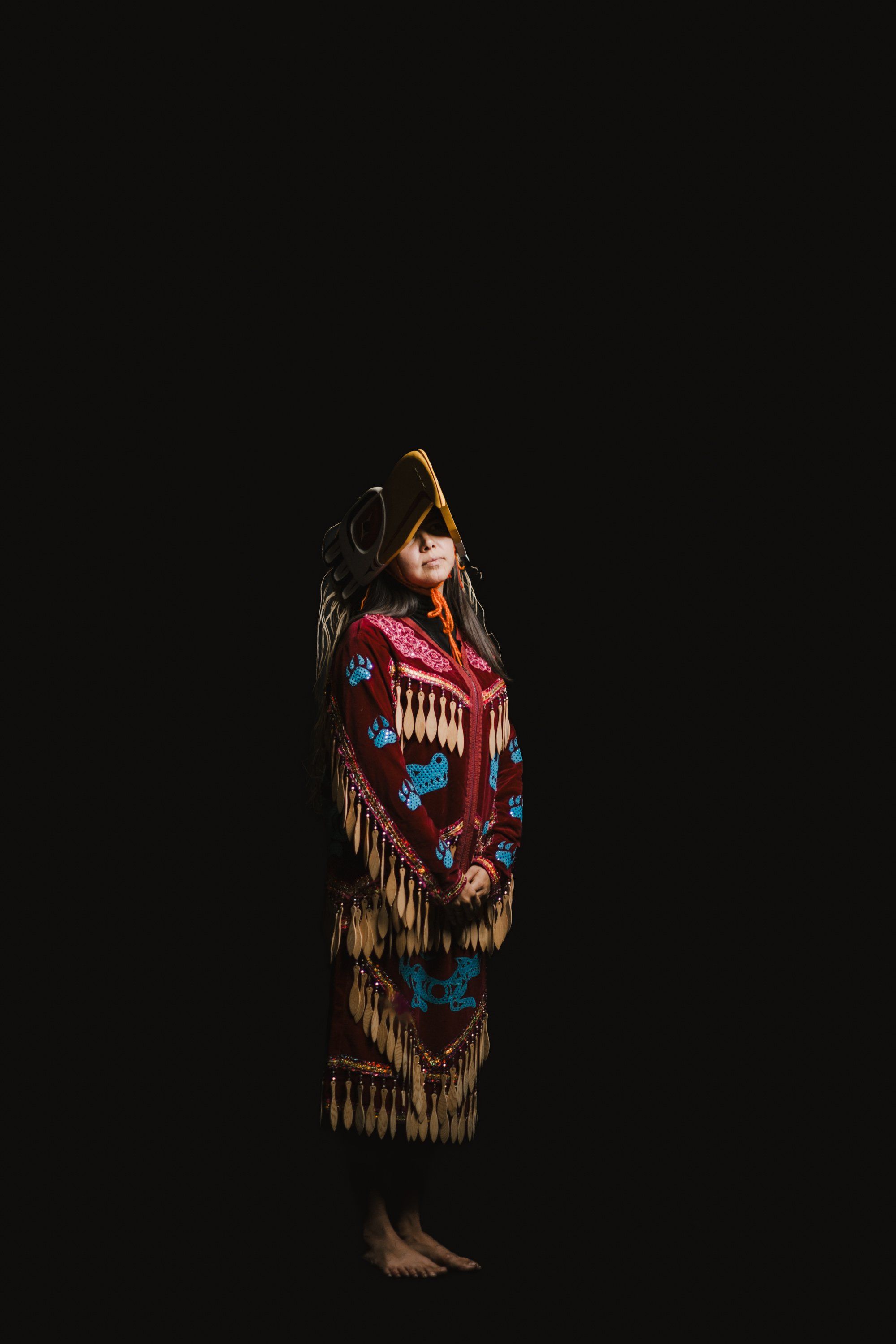 Woman in ornate red and blue regalia with fringe stands against black background, wearing traditional Coast Salish ceremonial dress