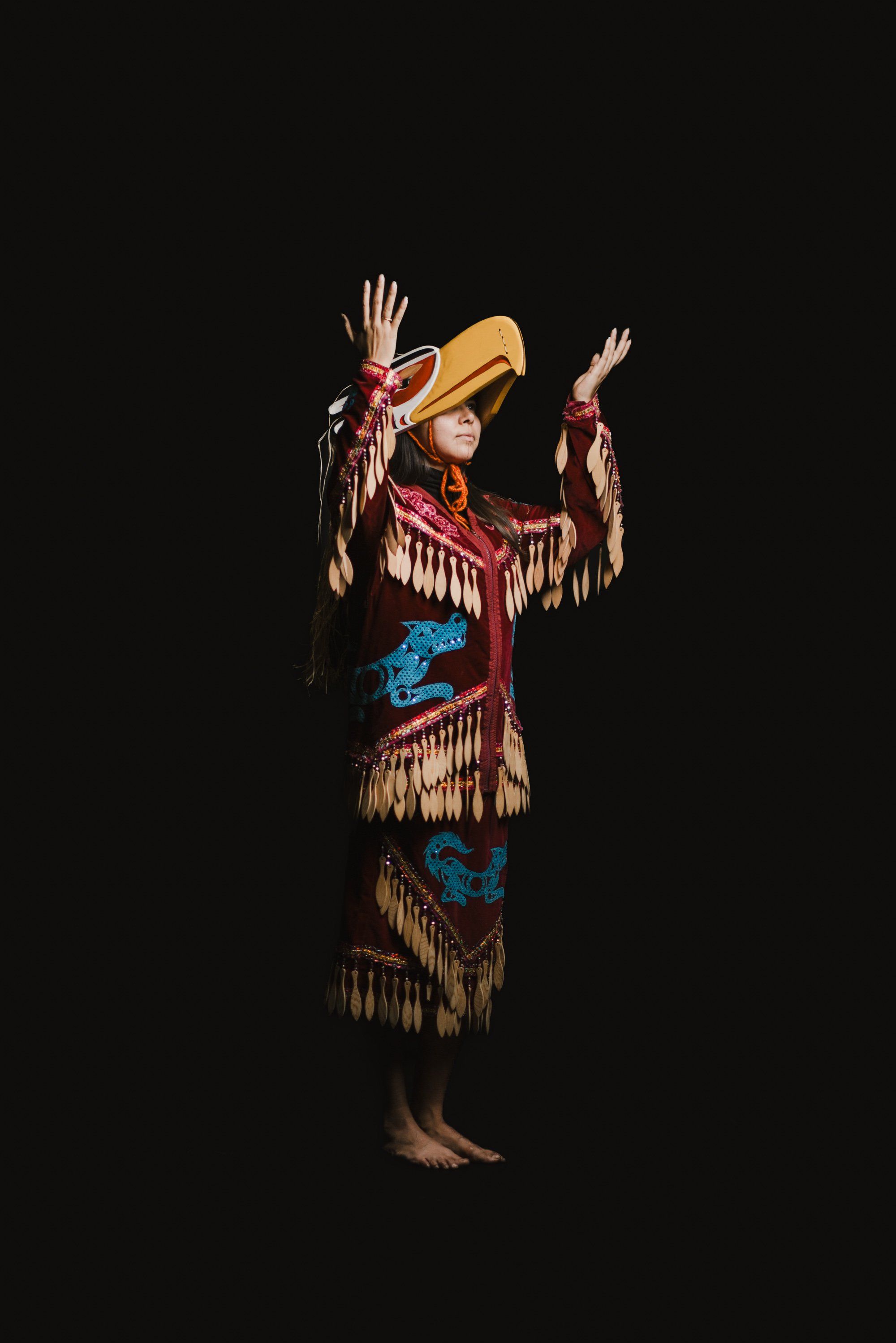 Dancer in traditional Coast Salish regalia with fringed leather dress and wide-brimmed hat raises arms against black background