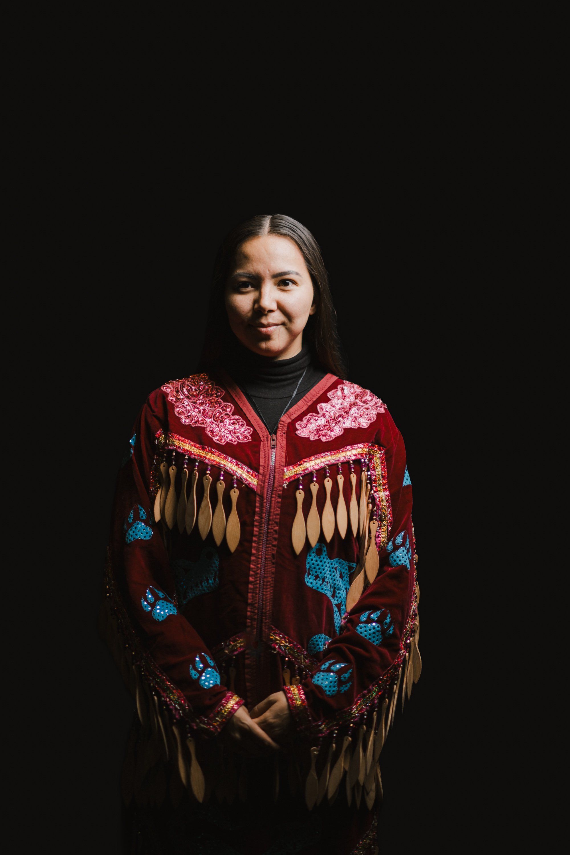 Woman in red and blue beaded regalia with fringe against black background, gentle smile, dramatic side lighting
