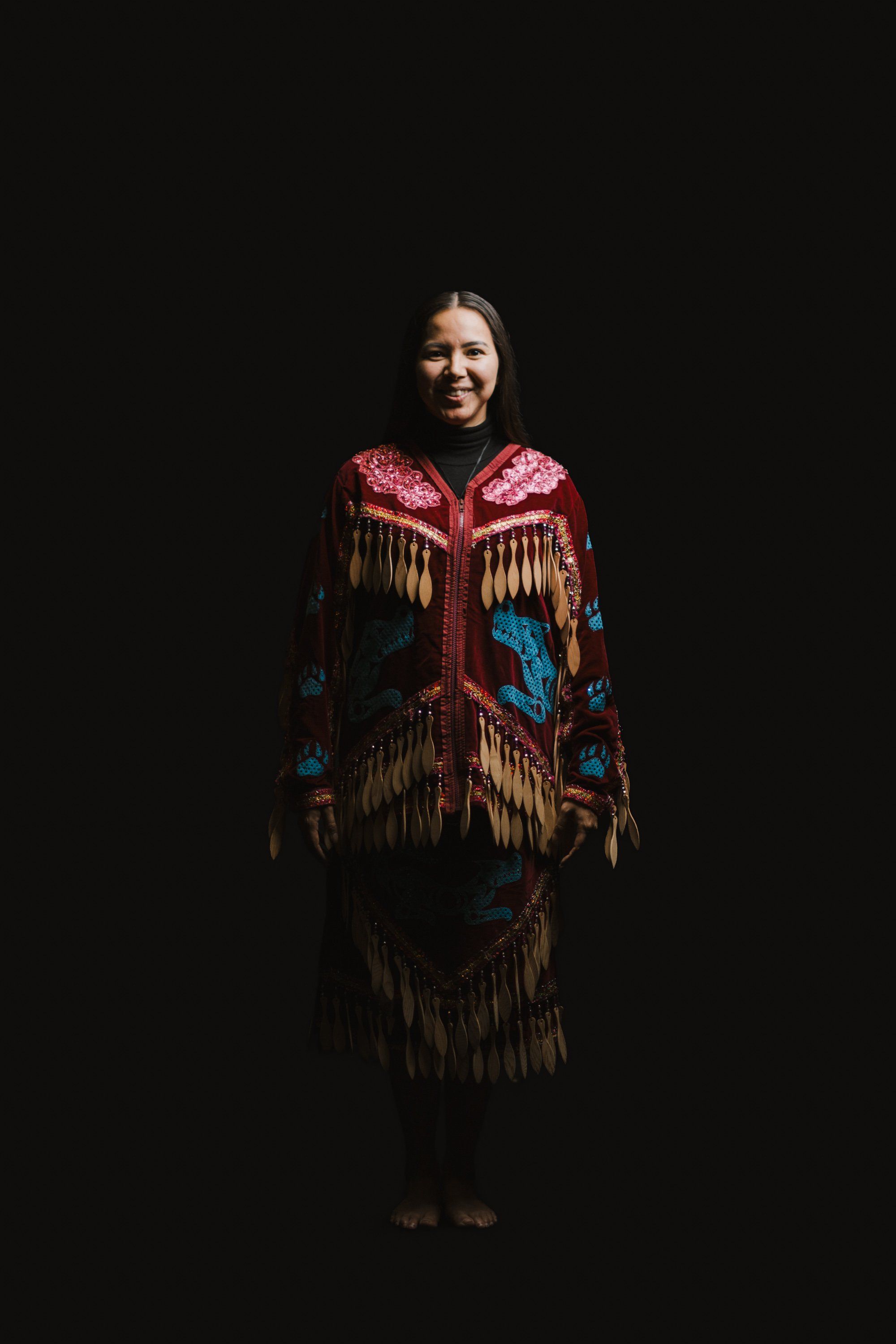 Smiling woman in ornate Coast Salish regalia with red and blue beadwork and fringe stands against black background