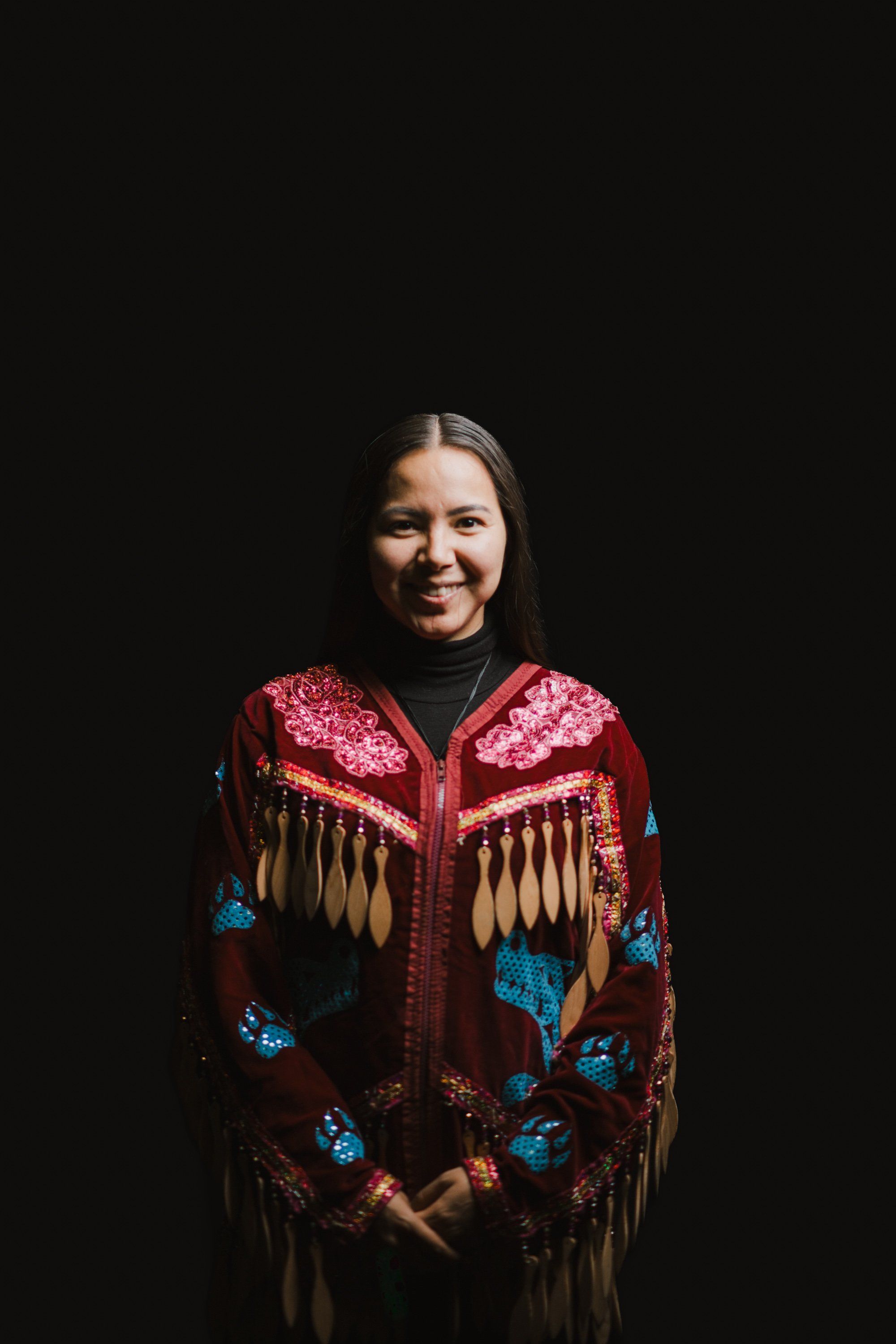 Woman in ornate red and blue beaded regalia with fringe against black background, smiling warmly at camera
