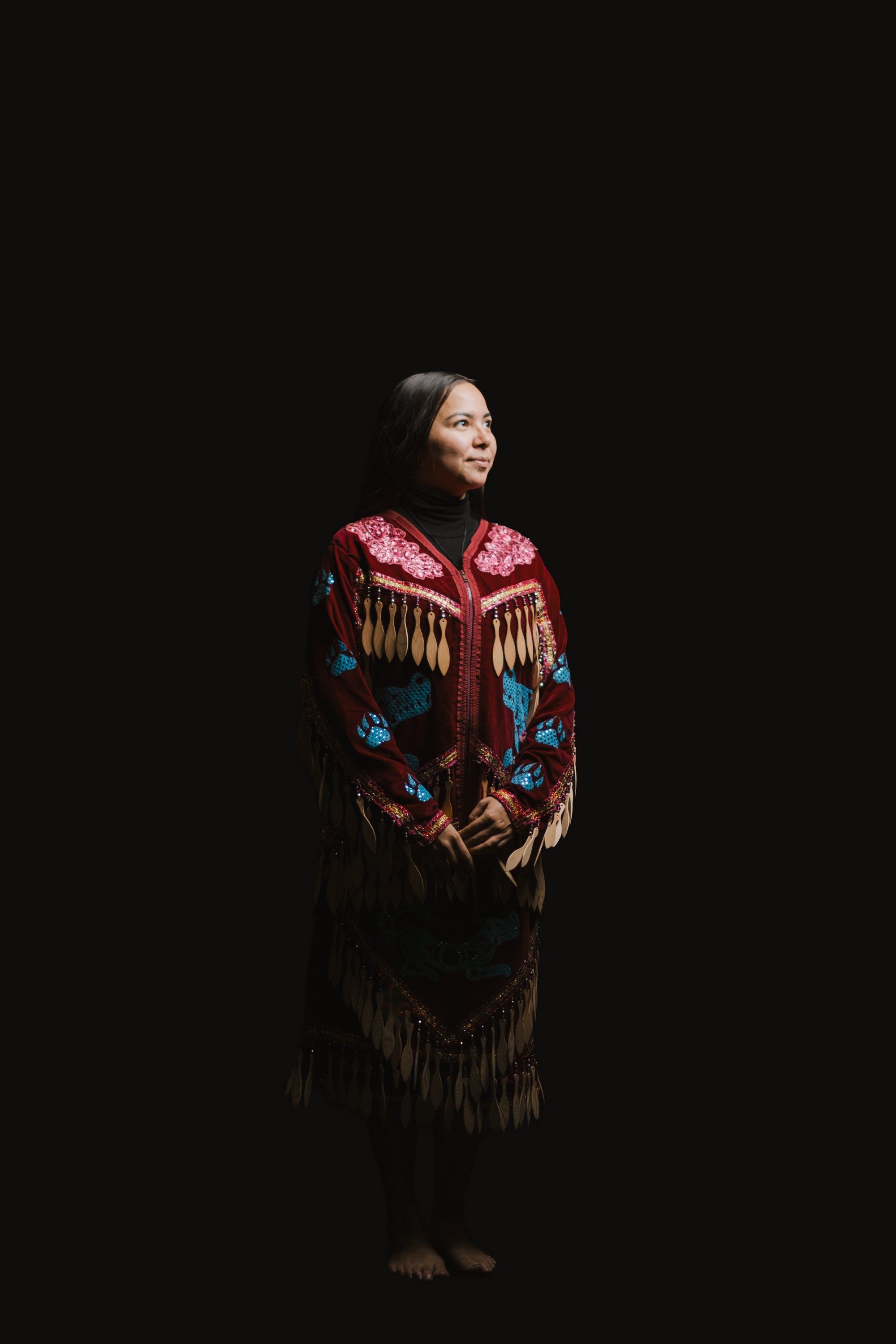 Woman in ornate red and blue beaded regalia with fringe stands against black background, gazing left in contemplative pose
