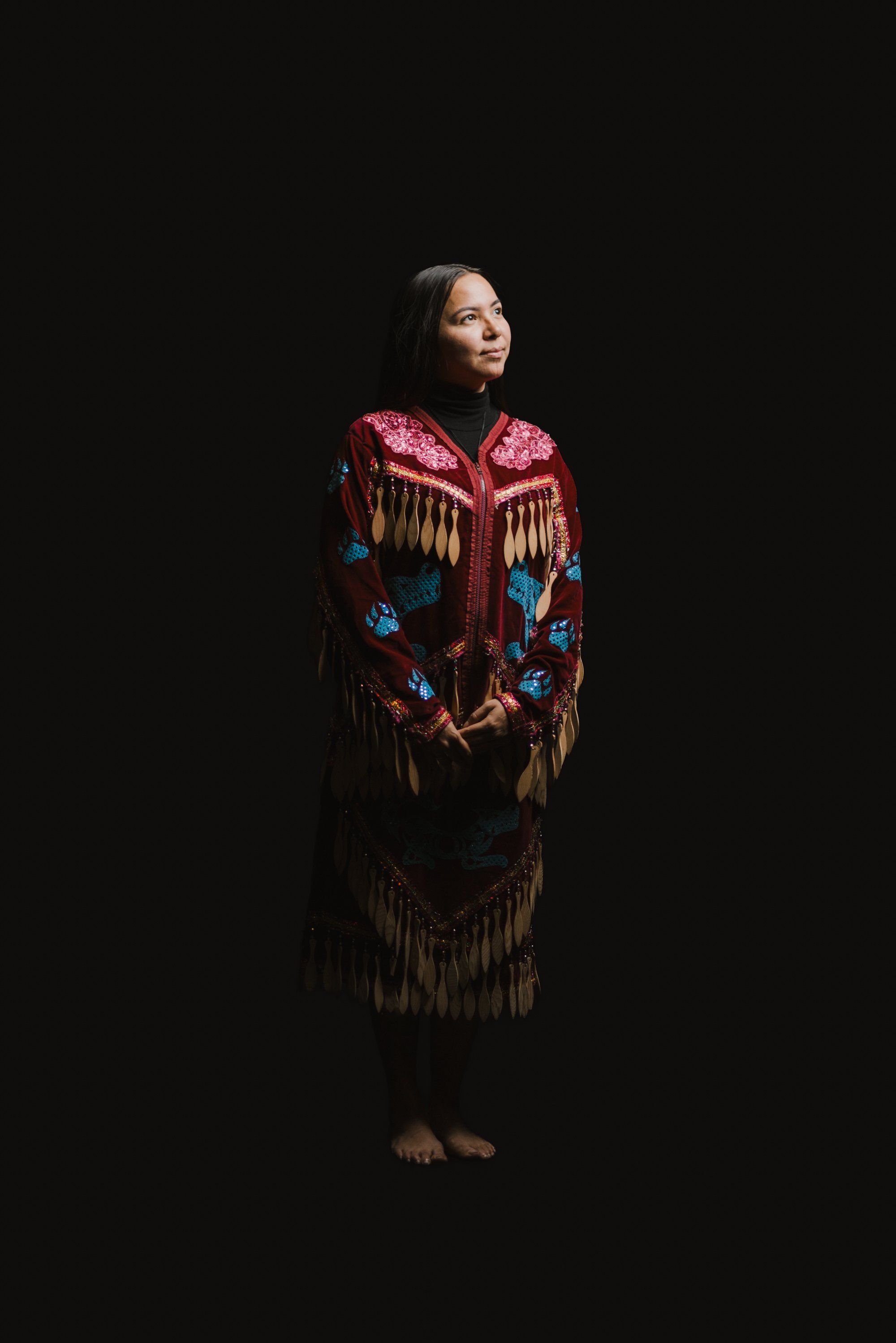 Woman in ornate red and black regalia with fringe and beadwork stands barefoot against black background, gazing upward