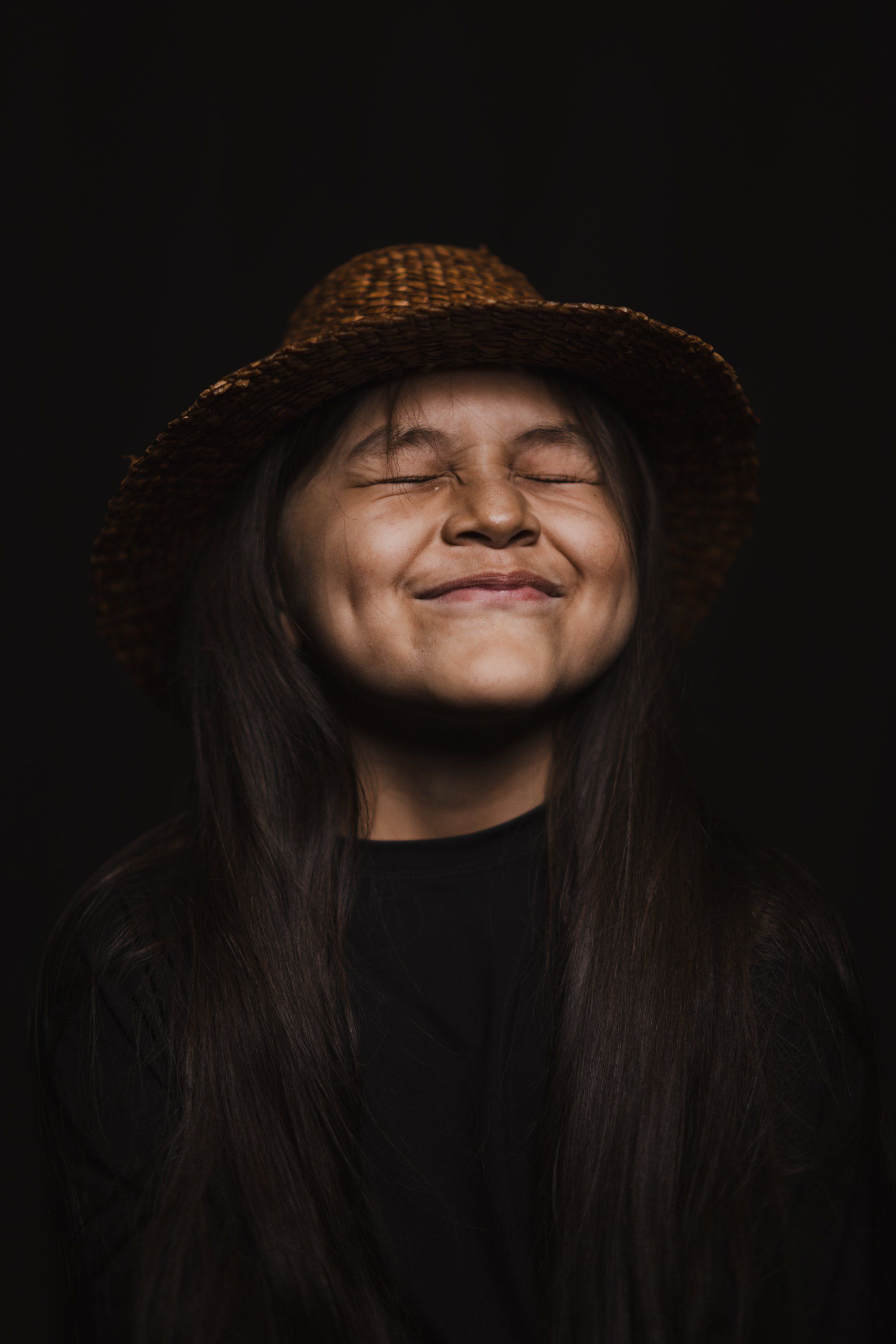 Person with long dark hair and closed eyes wearing a woven hat, smiling warmly against a dark background