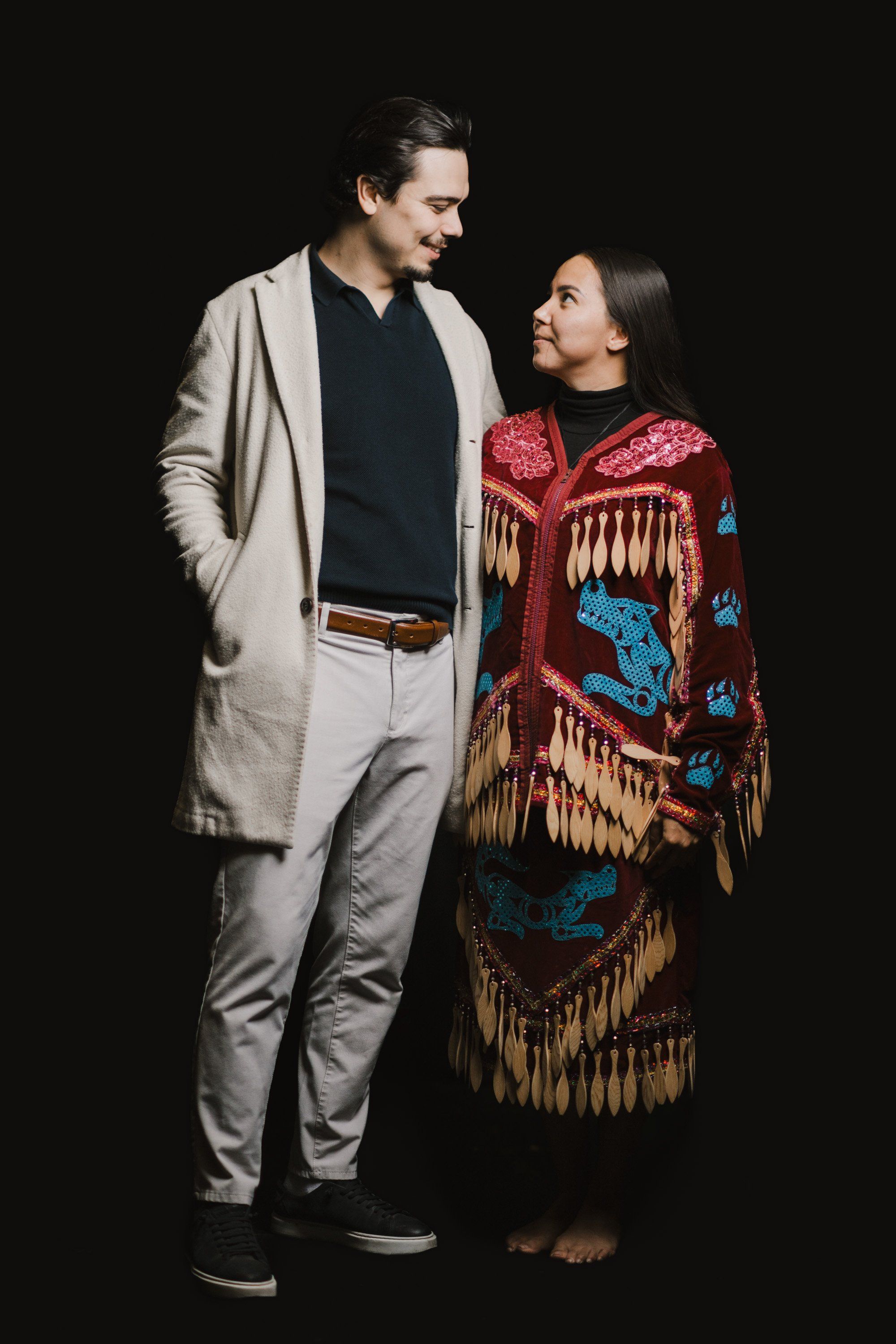 Man in cream cardigan and woman in red Coast Salish regalia stand together against black background, looking toward each other with warm intimacy