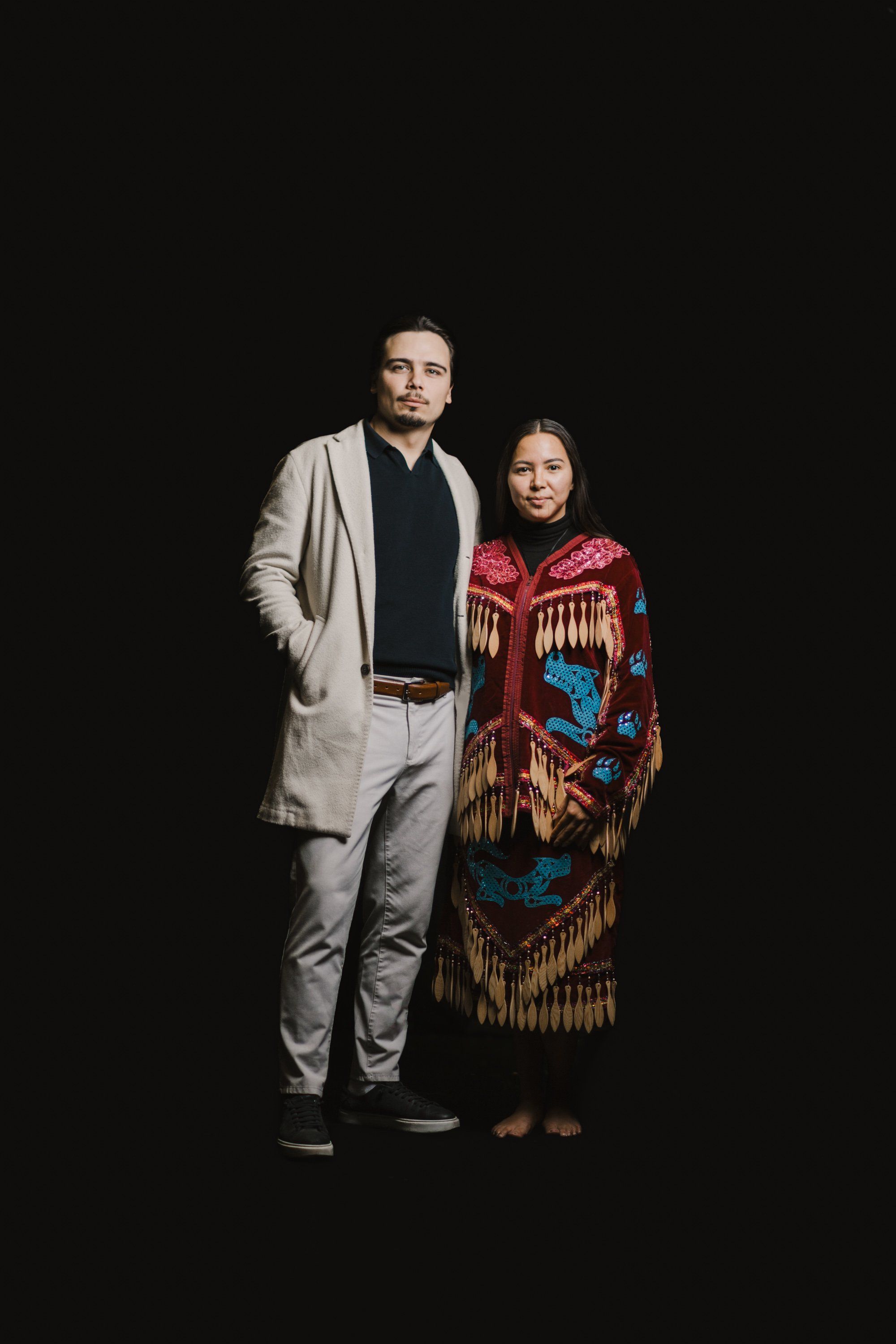Man in light grey blazer and woman in red and gold Coast Salish regalia stand together against black background, studio portrait lighting