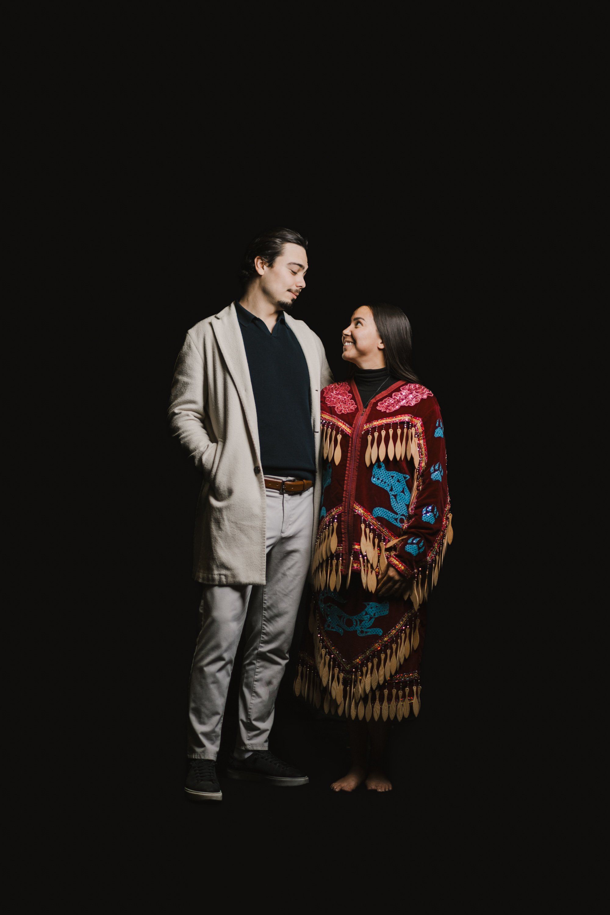 Man in gray blazer and woman in red and gold Coast Salish regalia face each other against black background, intimate portrait lighting