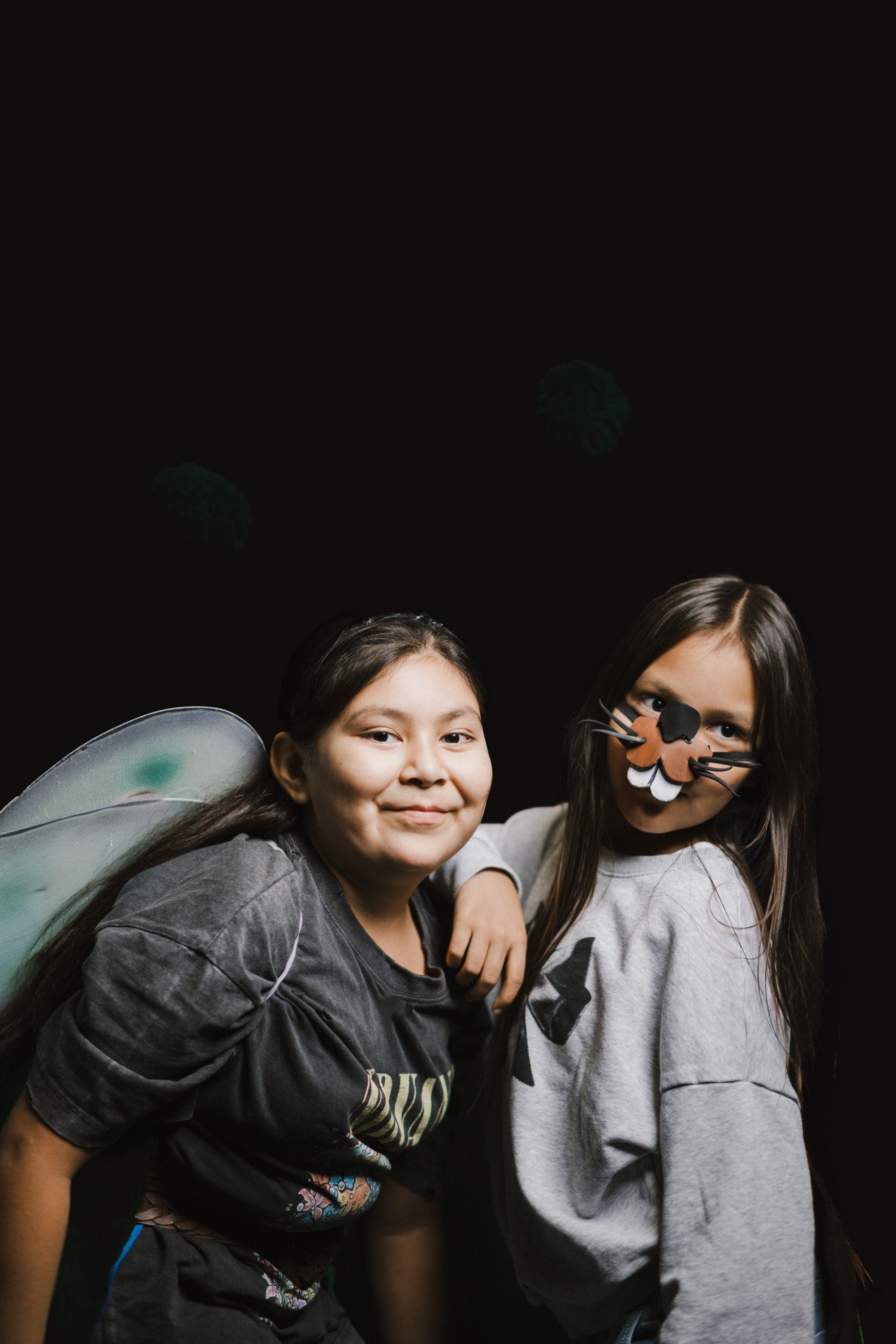 Two young people smile at the camera against a black background, one wearing glasses and a gray shirt, the other in a dark shirt with a blue-green feathered…