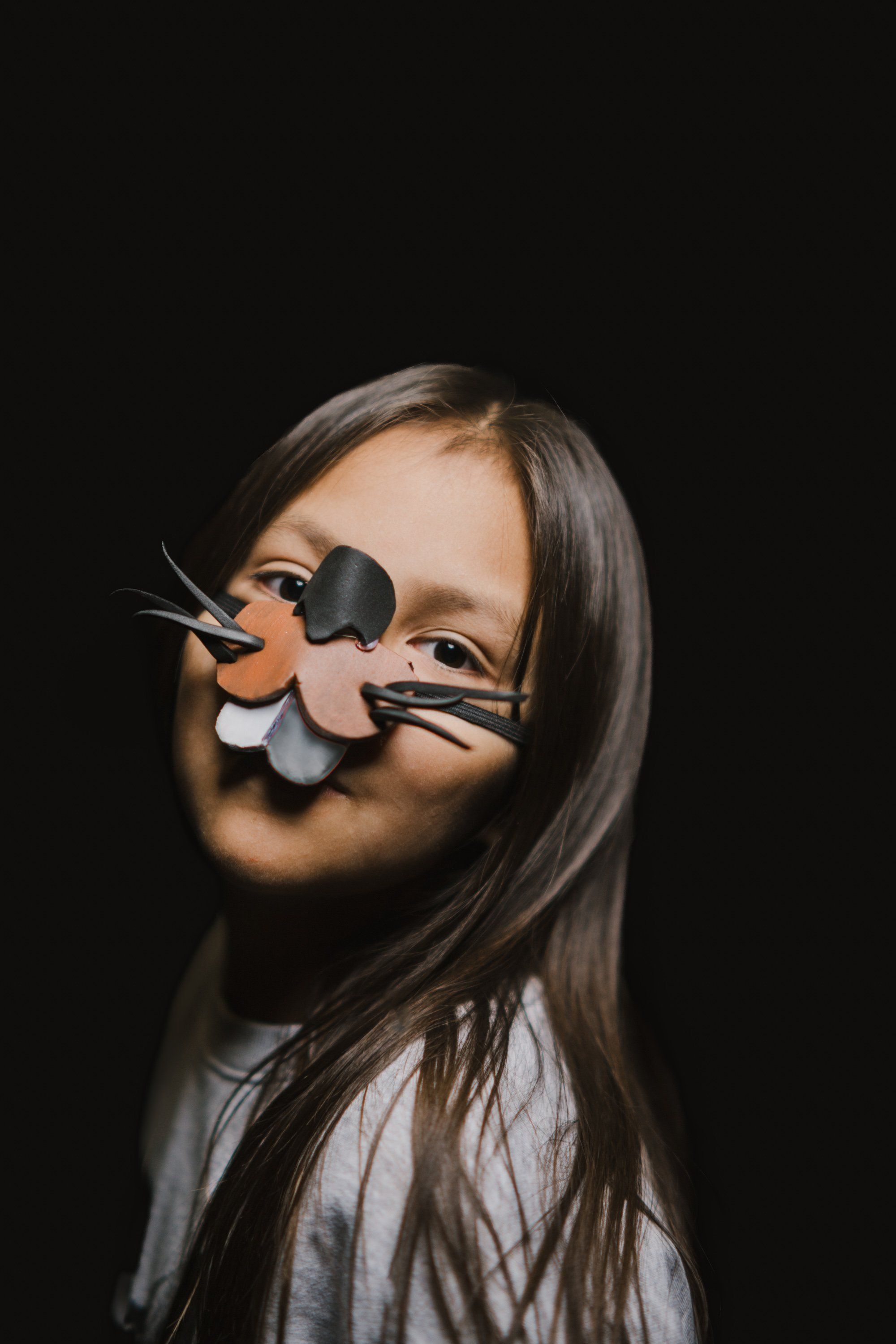 Young girl with long dark hair wearing an elaborate carved wooden mask against a black background, smiling warmly