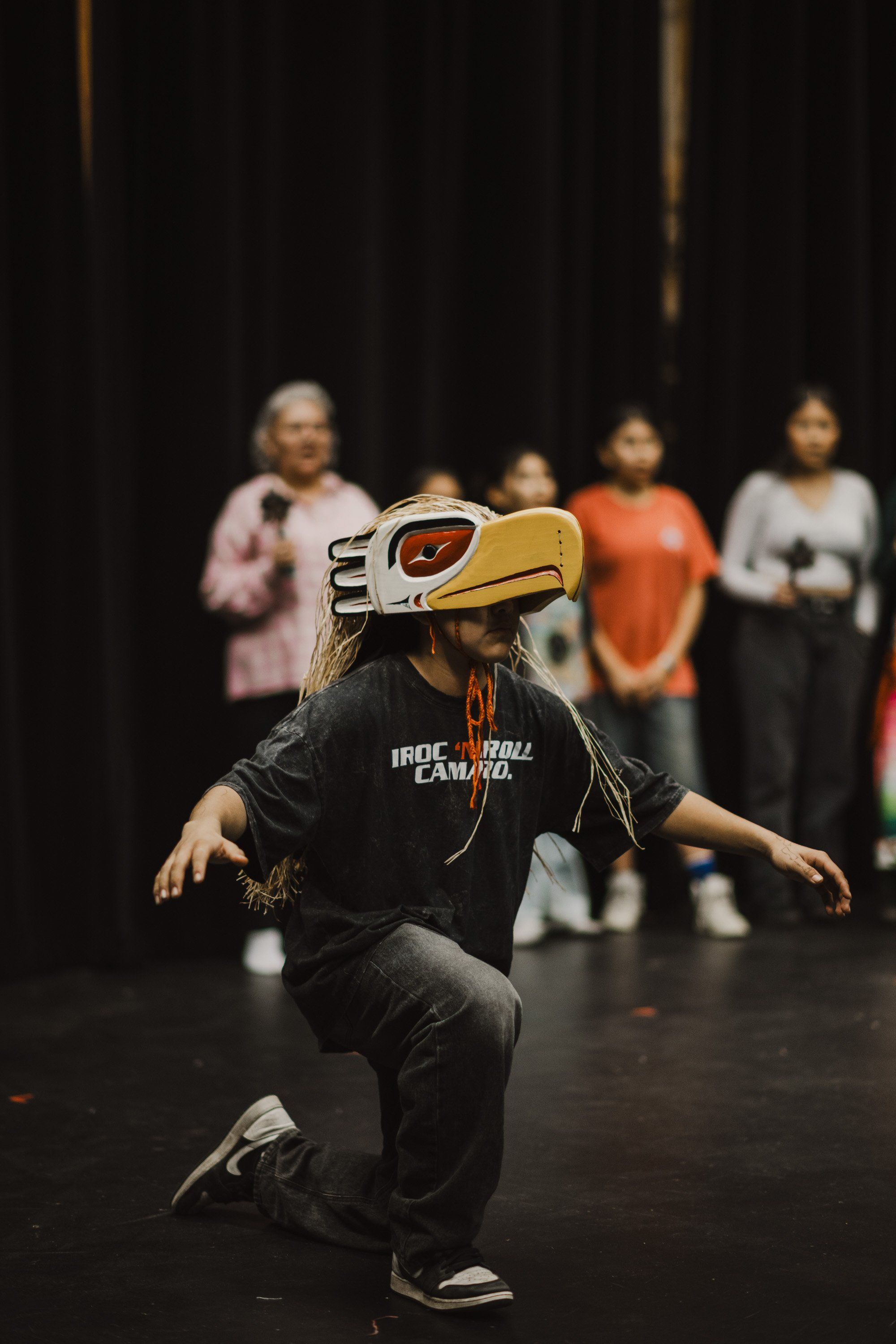 Child in golden eagle mask dances on dark stage while spectators watch from backdrop, celebrating Coast Salish culture