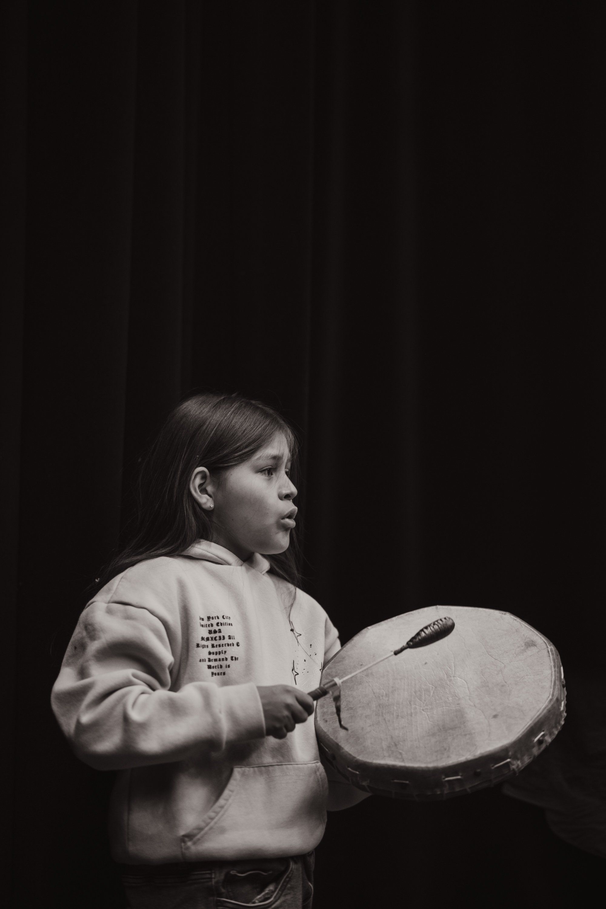 Young performer in profile holding a large drum against a dark backdrop, wearing a light-colored sweatshirt with text detail