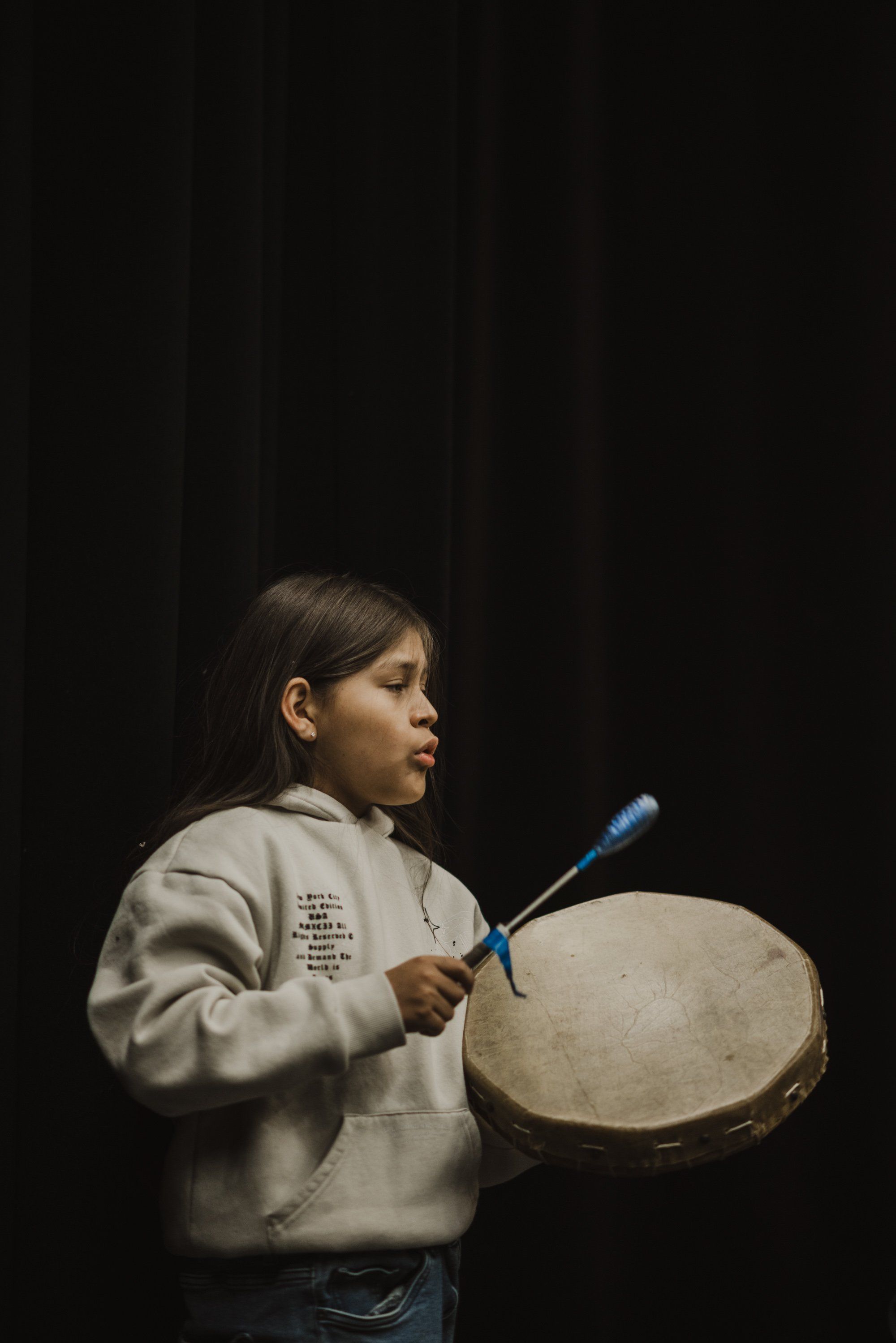 Young musician in profile holding a large traditional drum with beater against dark background