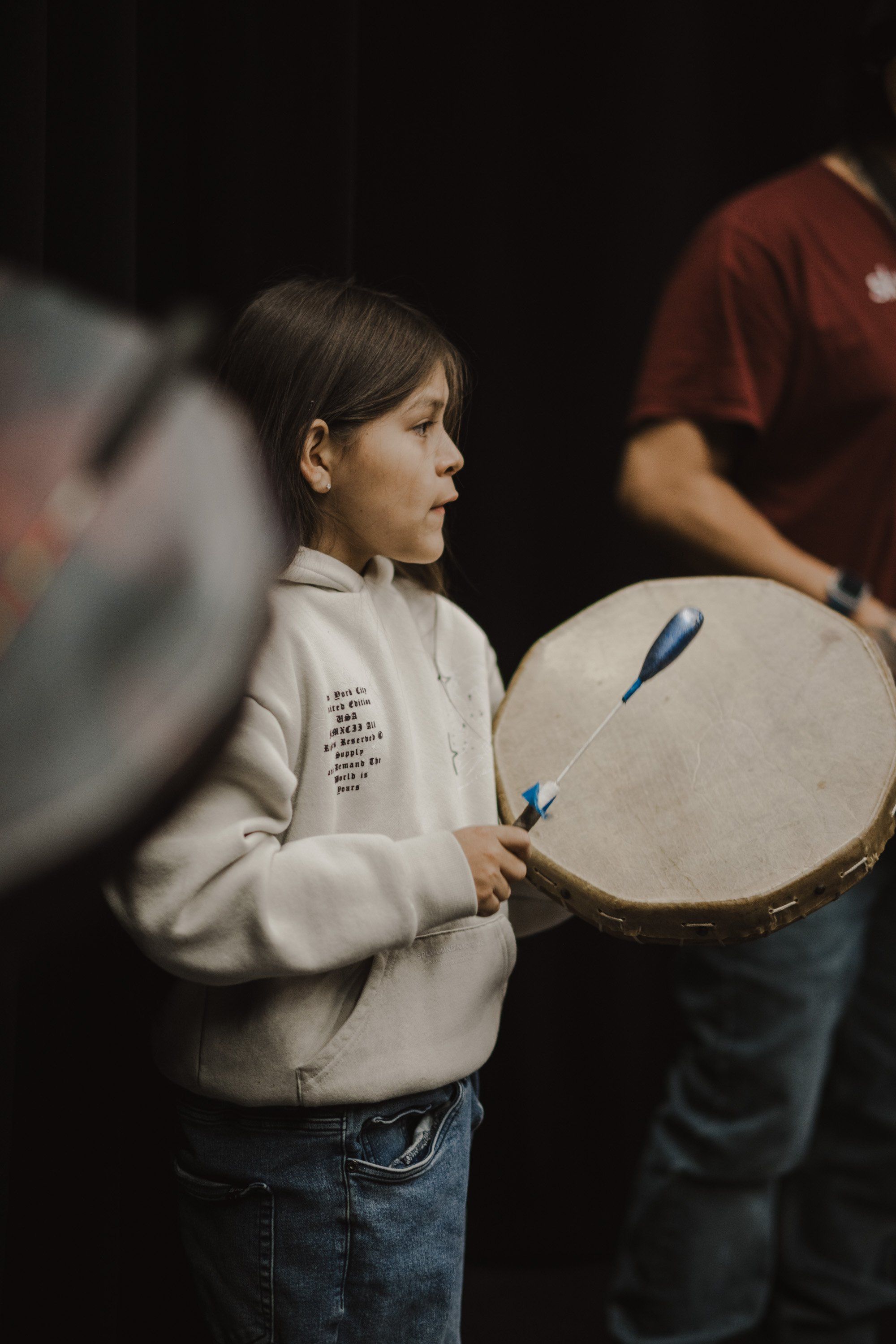 Young performer holding a large drum with blue drumstick, focused expression, dark background with blurred figures nearby