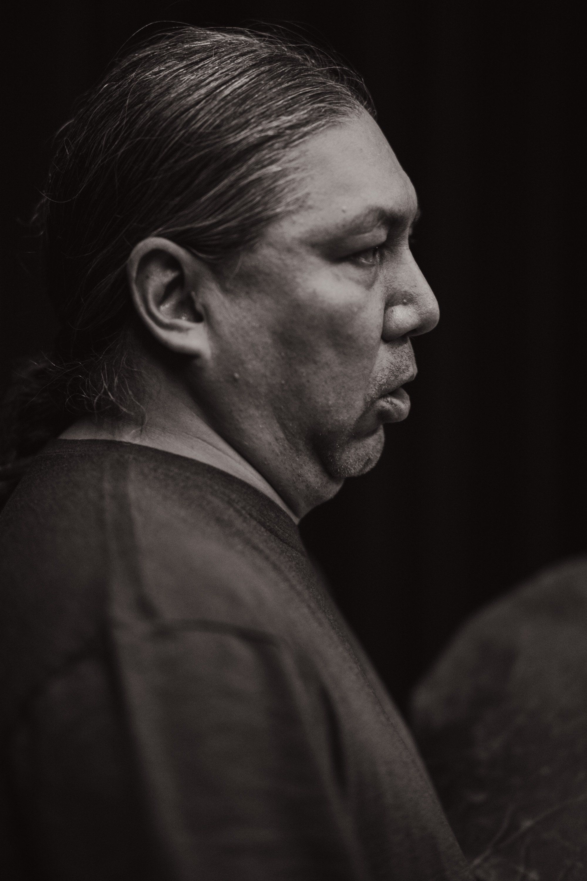 Profile portrait of a man with long dark hair against a black background, captured in dramatic side lighting that emphasizes facial features and contemplativ…
