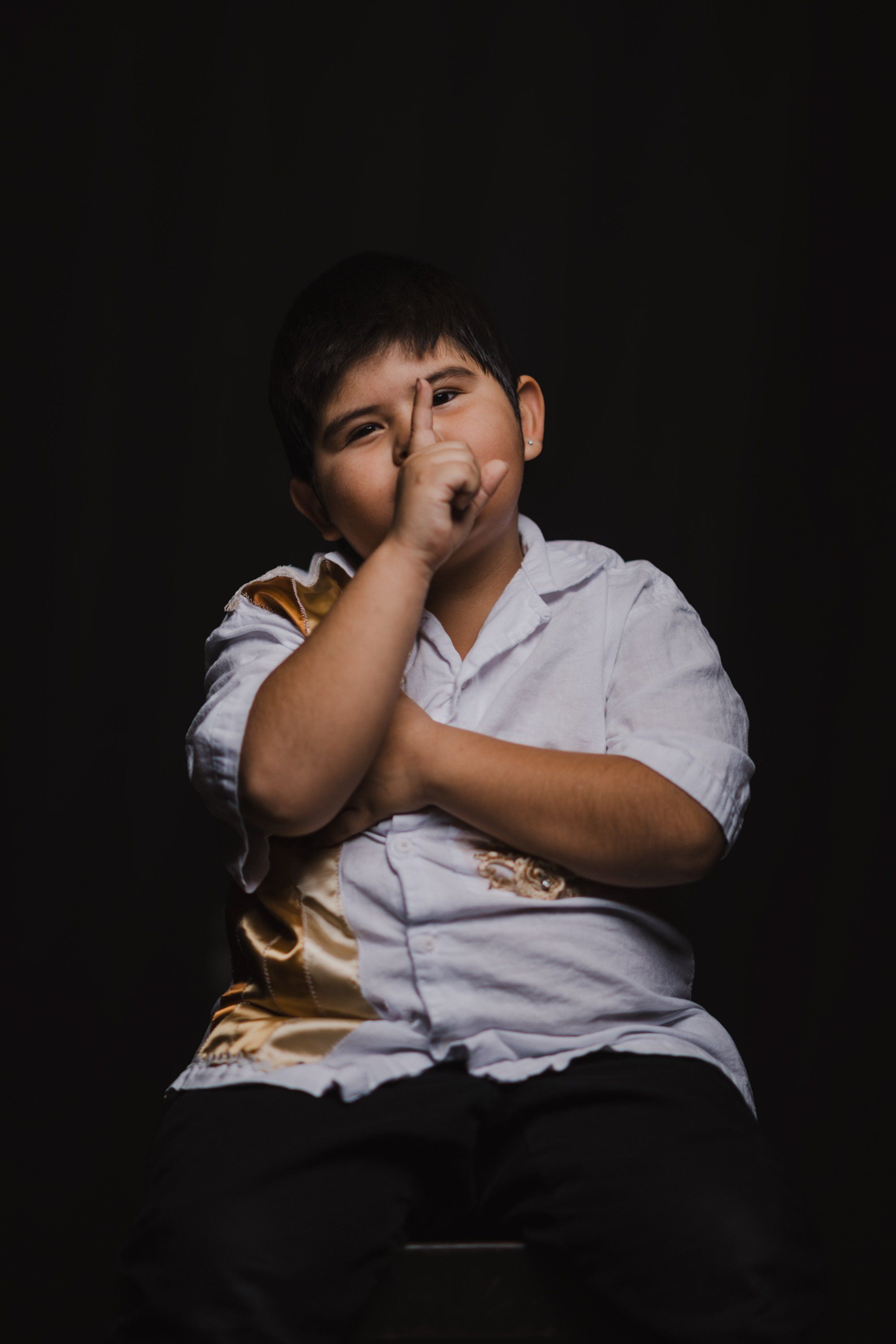 Young person in light blue shirt seated against black background, chin resting on hand in thoughtful pose, warm side lighting