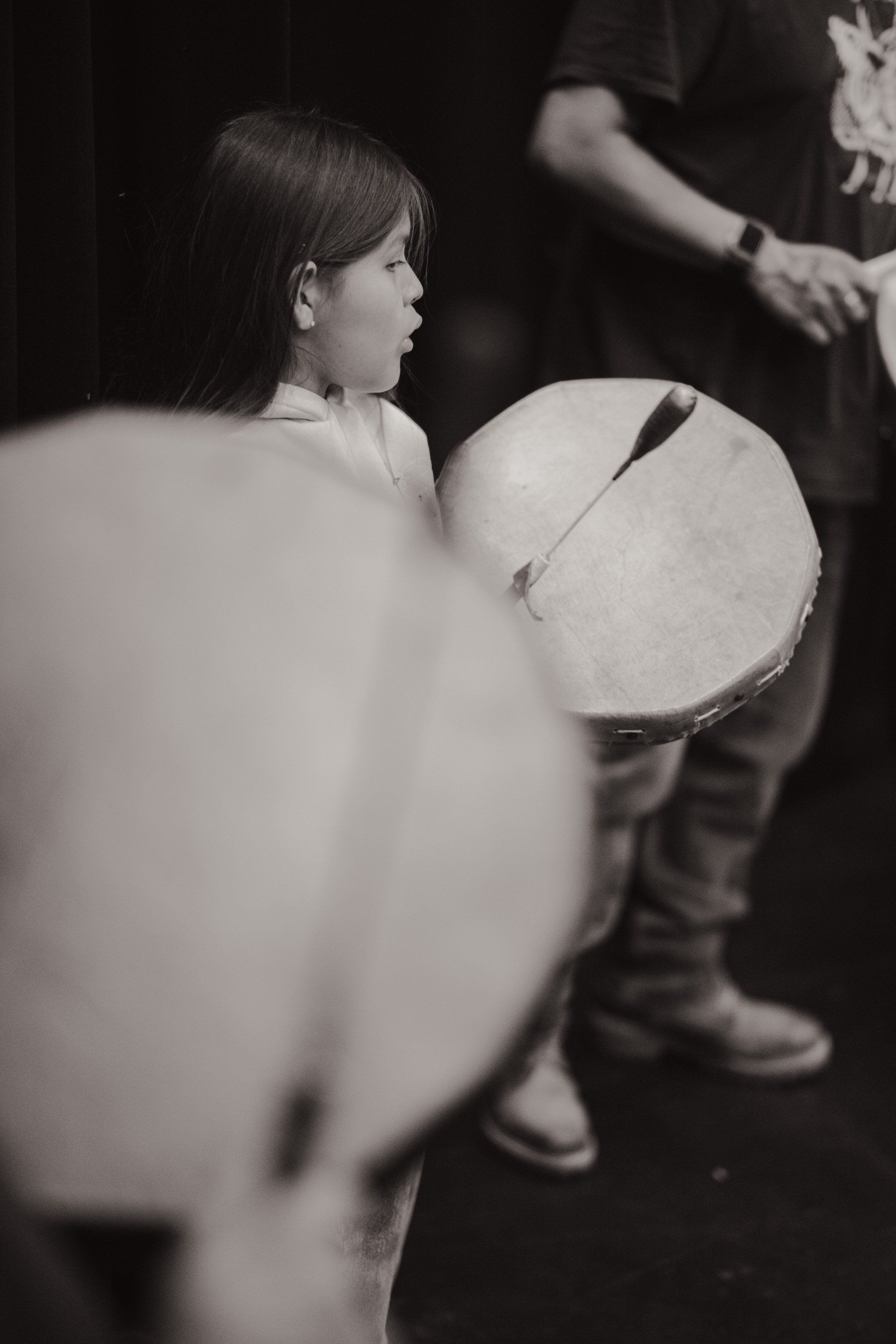 Young child in profile holding a large drum at a Coast Salish cultural celebration, dark background