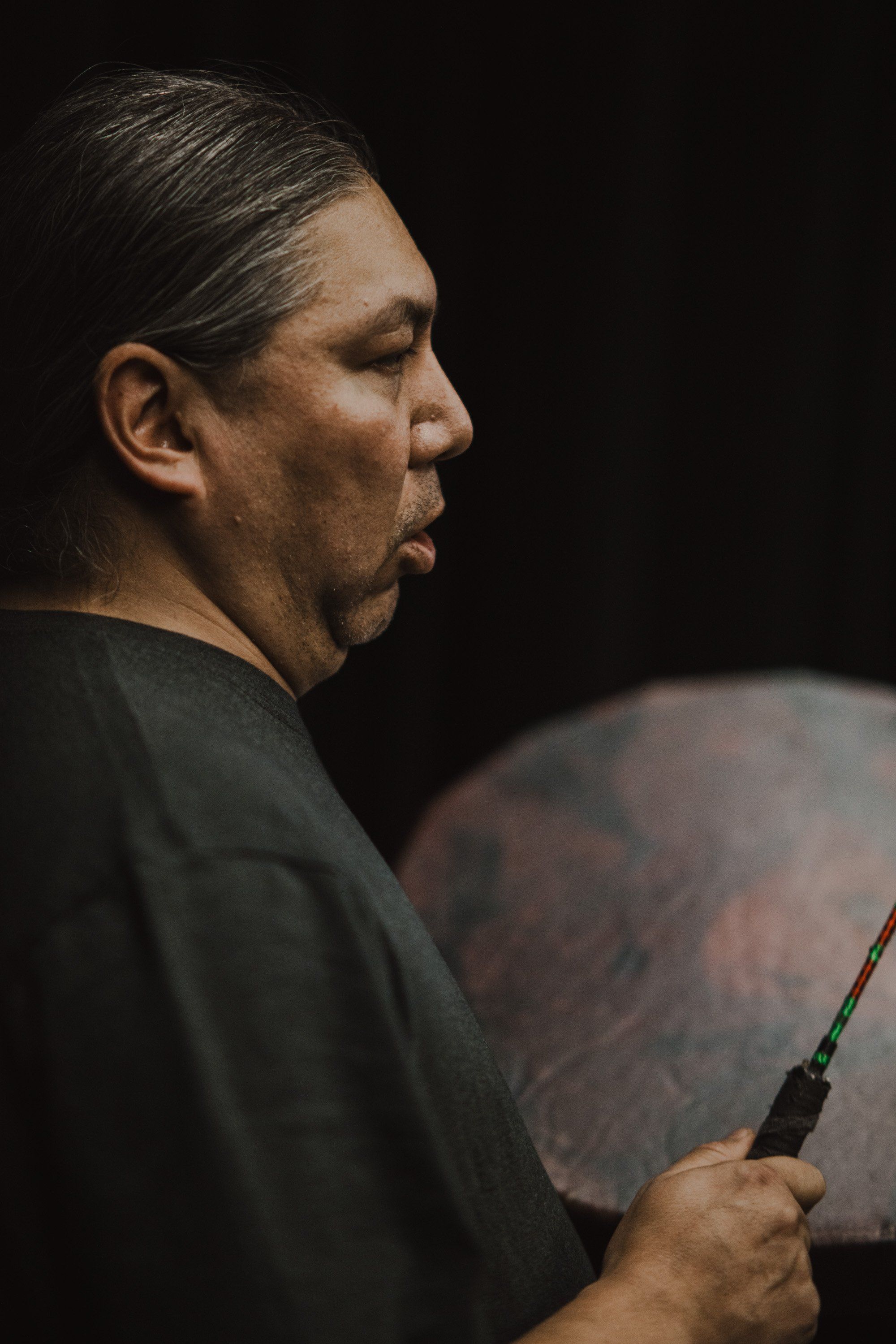 Coast Salish performer in profile holding a traditional drum, illuminated against black background during cultural celebration