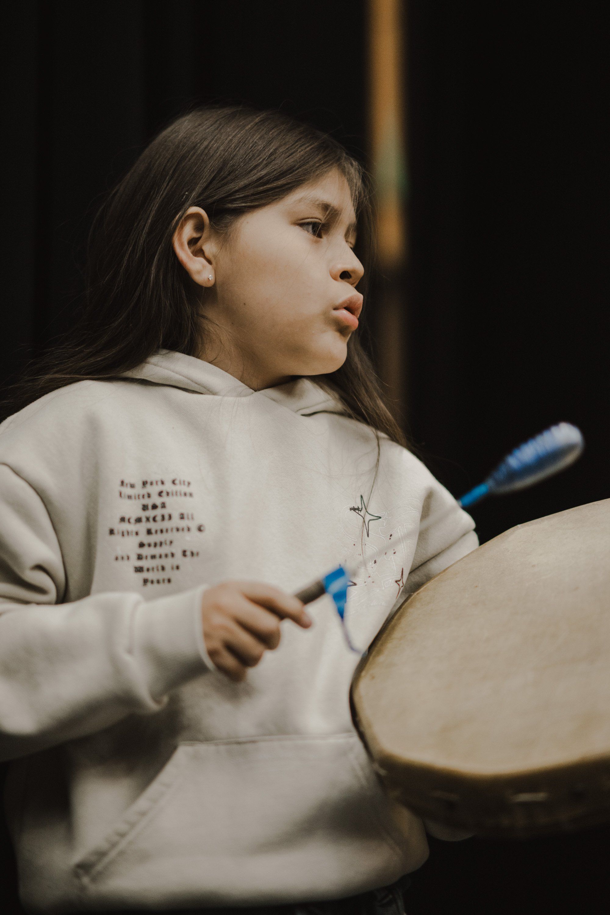 Young Coast Salish dancer in beige hoodie holds blue drumsticks over a large drum, captured in profile against dark background