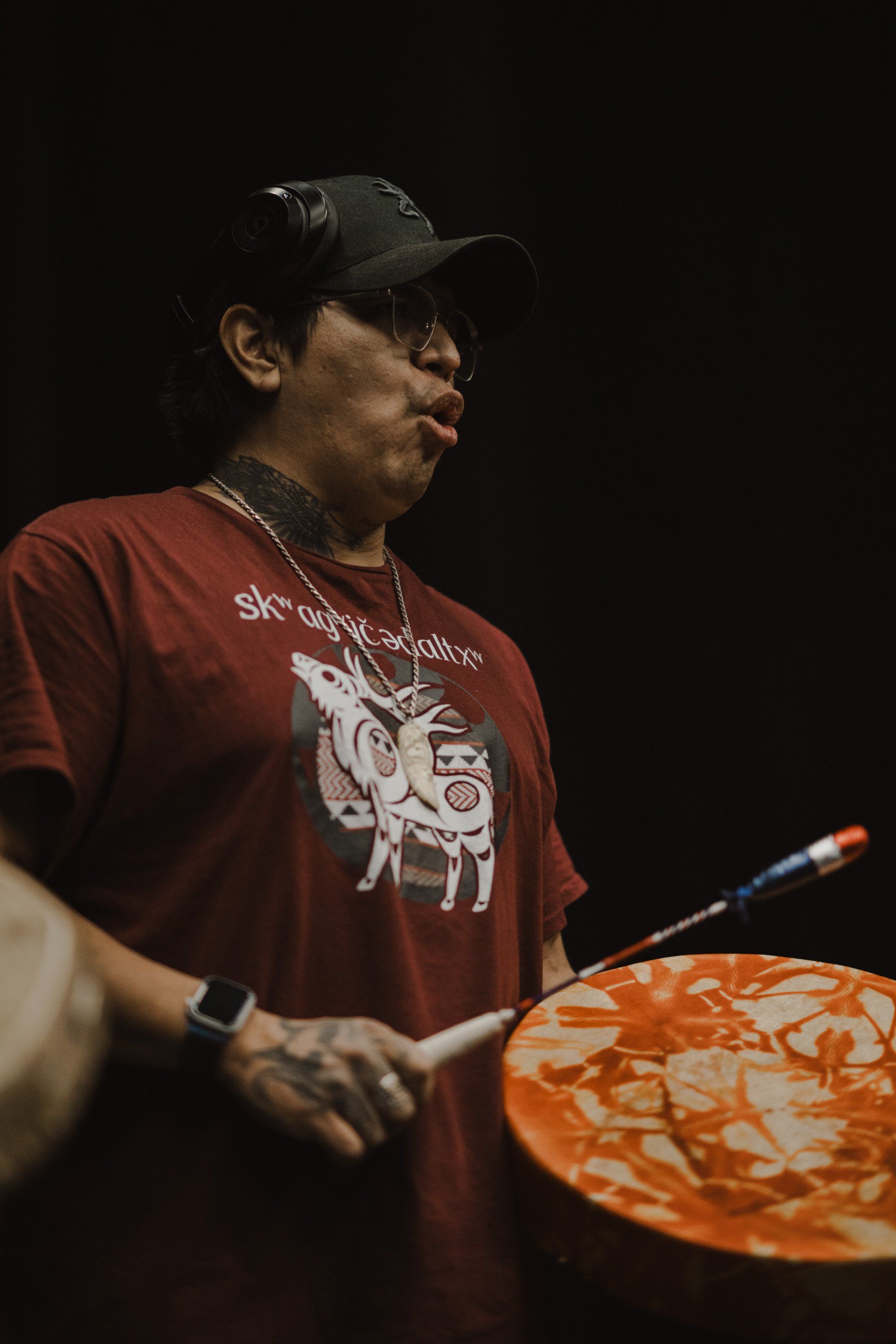 Performer in burgundy graphic tee and cap holds decorated drum against dark background, captured in profile during Coast Salish cultural presentation