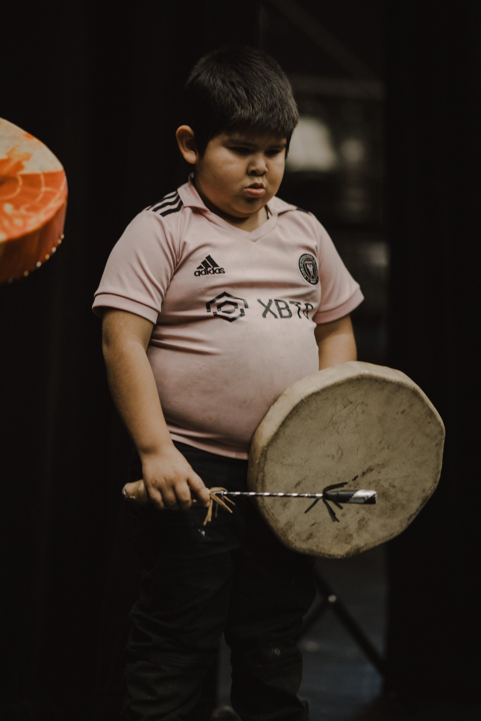 Young child in pink Adidas shirt concentrating while holding a traditional drum against dark background