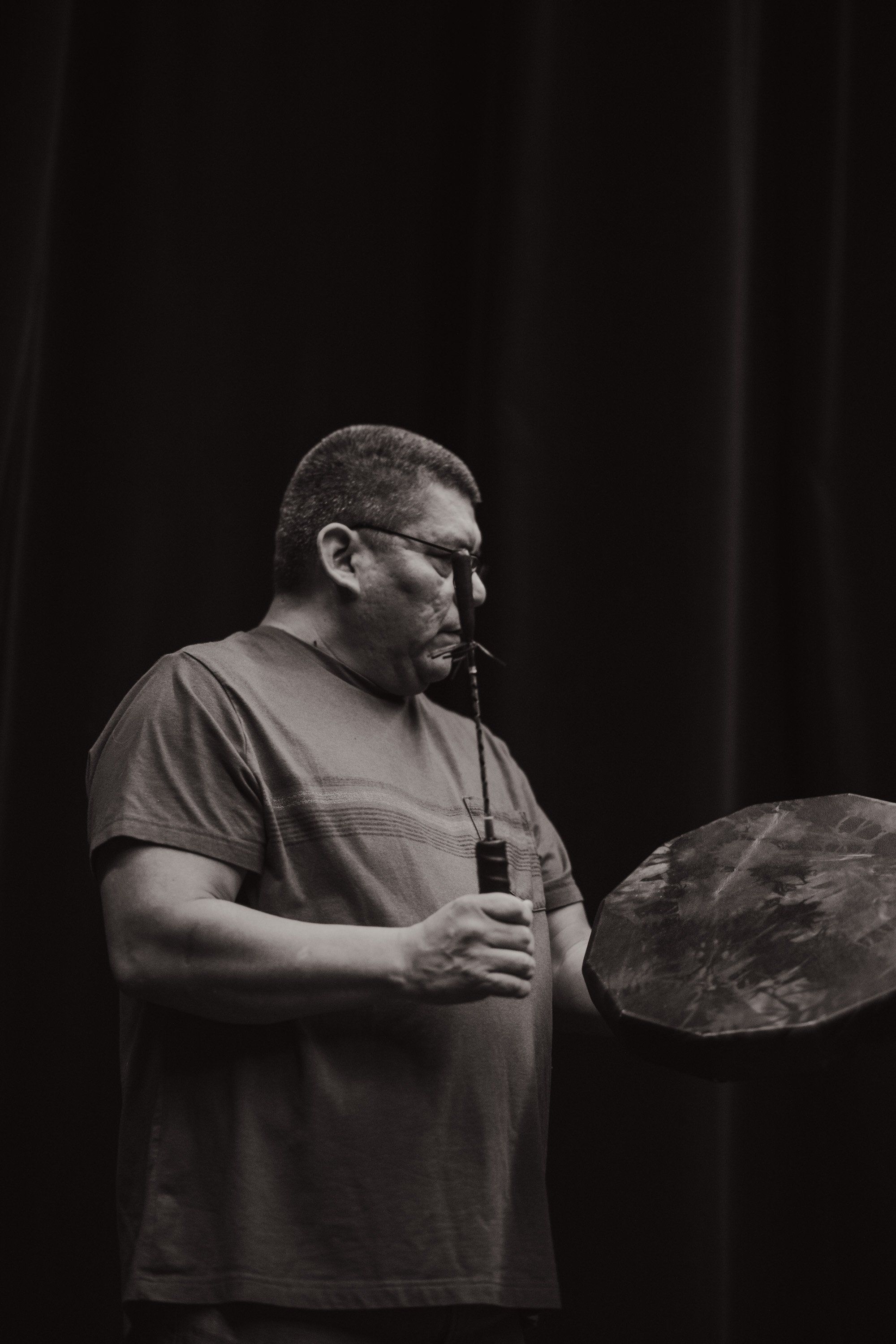 Man in glasses speaking into microphone against dark backdrop, holding what appears to be a traditional Indigenous artwork or object