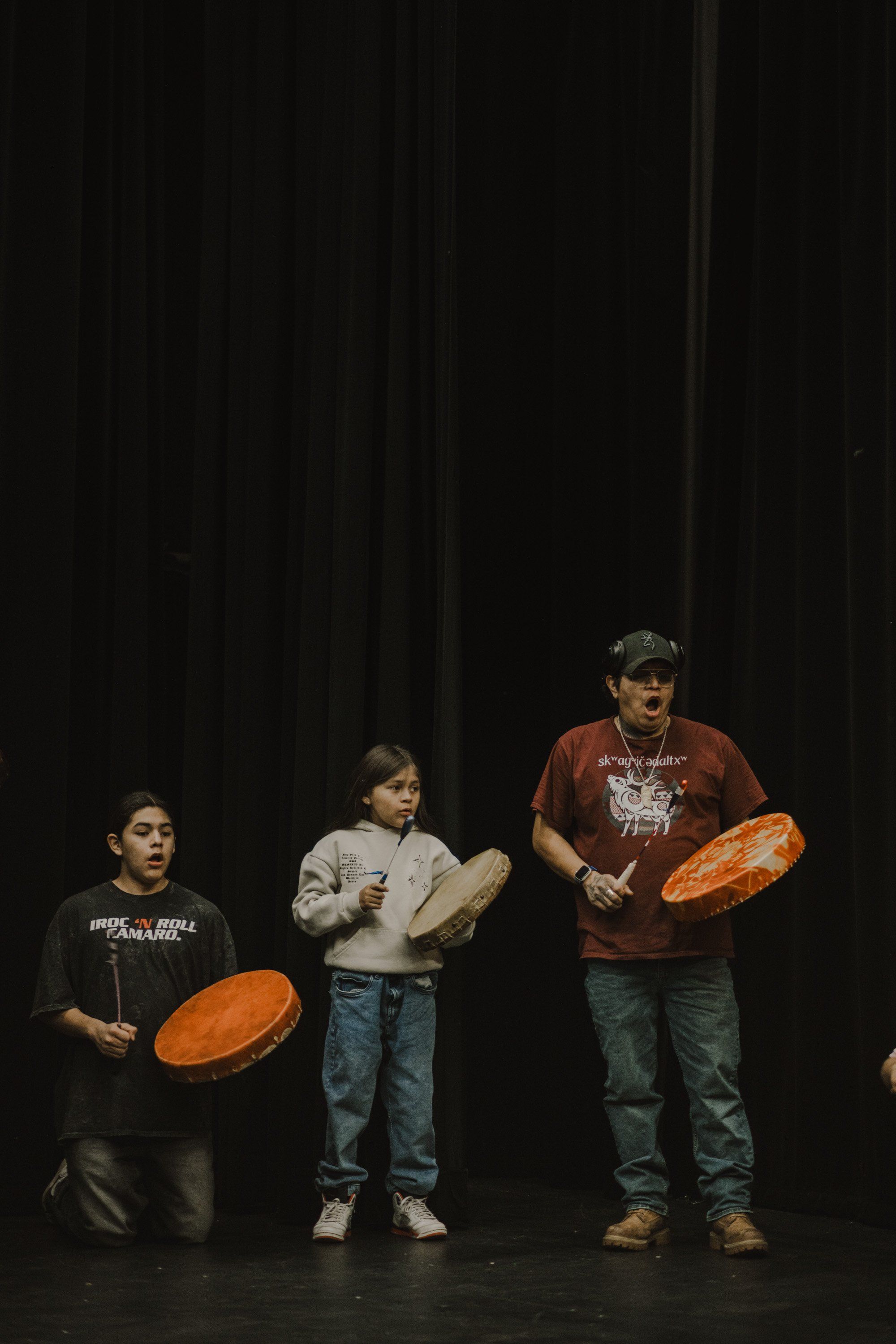 Three young performers stand against a black curtain holding orange bowls, wearing casual clothing and caps in a staged performance setting