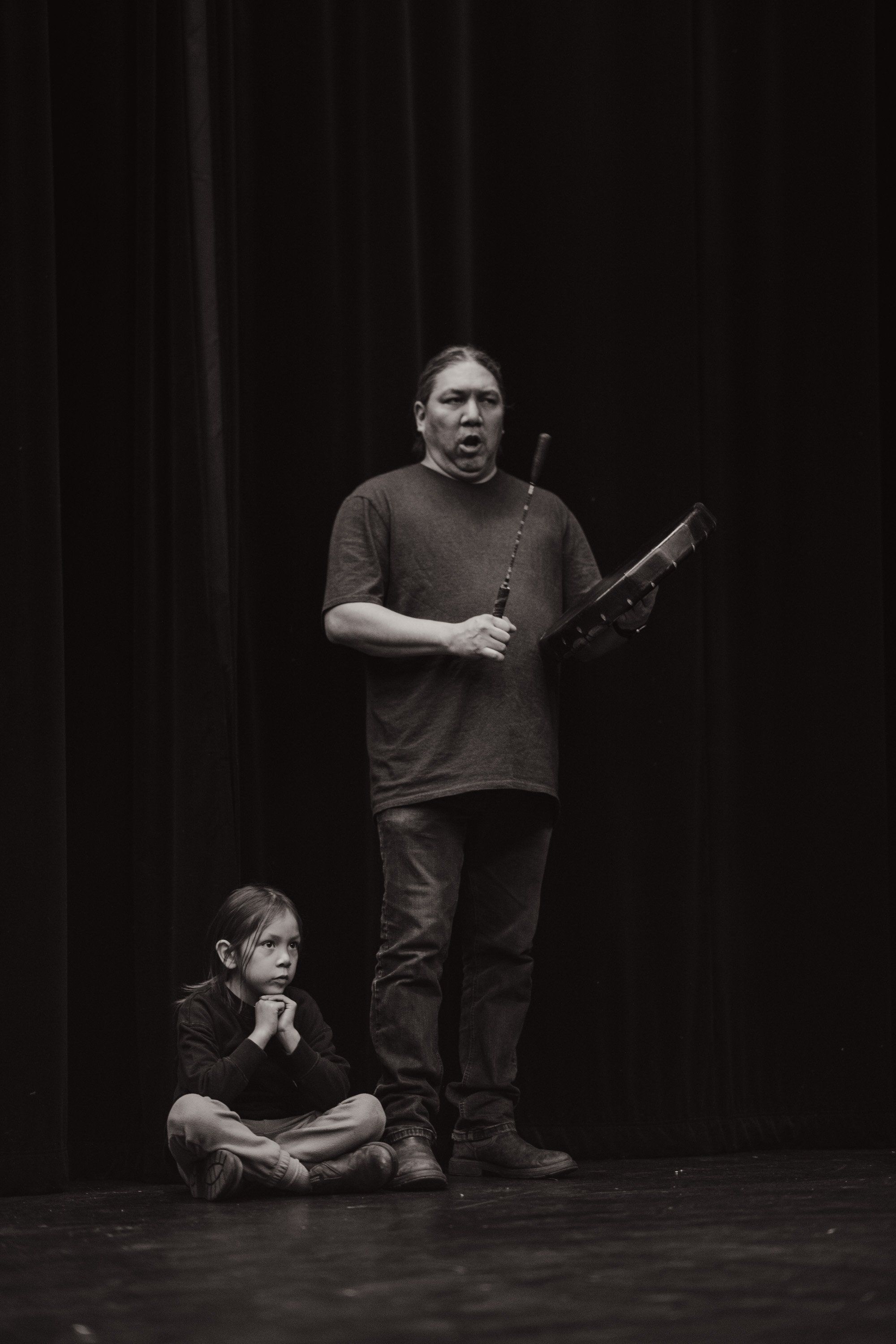 Adult and child on dark stage, adult gesturing expressively while holding carved wooden object, child seated and listening intently