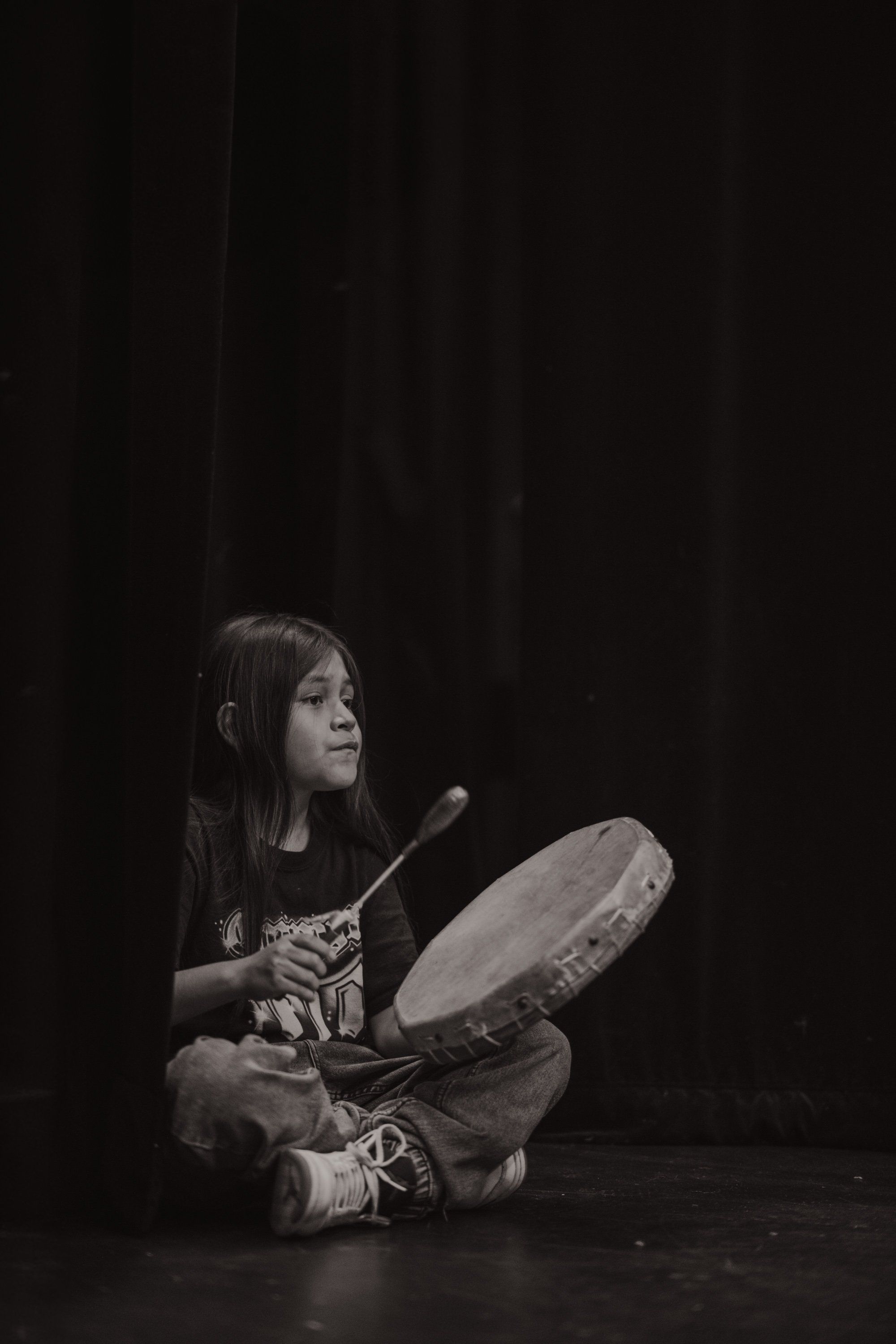 Young performer seated on dark stage holding a large drum, gazing upward in contemplative expression during Coast Salish cultural celebration