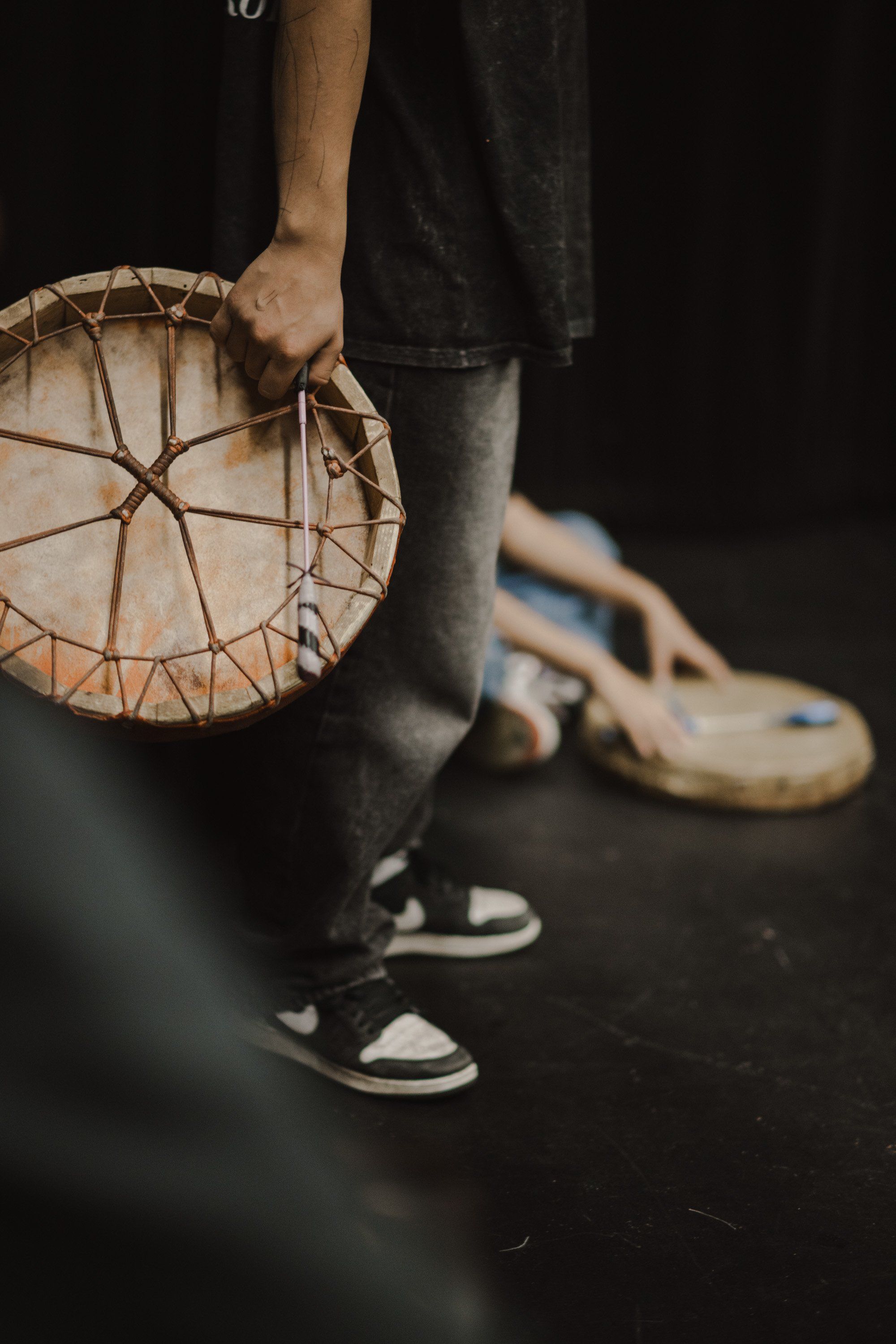 Person in dark clothing holds a circular woven basket while wearing white sneakers against a black background
