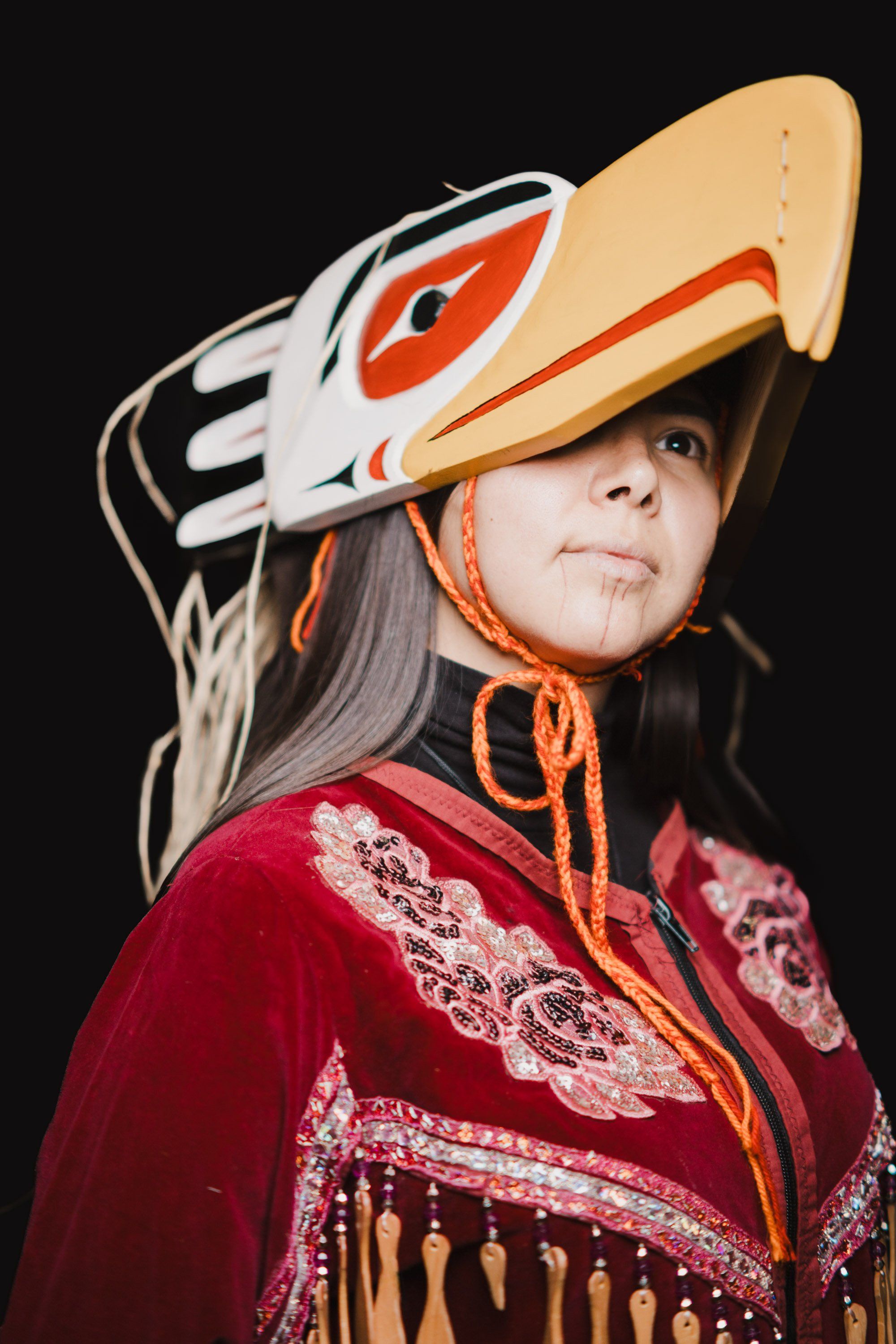 Elder in traditional Coast Salish regalia with carved wolf headdress and red ceremonial blanket against black background