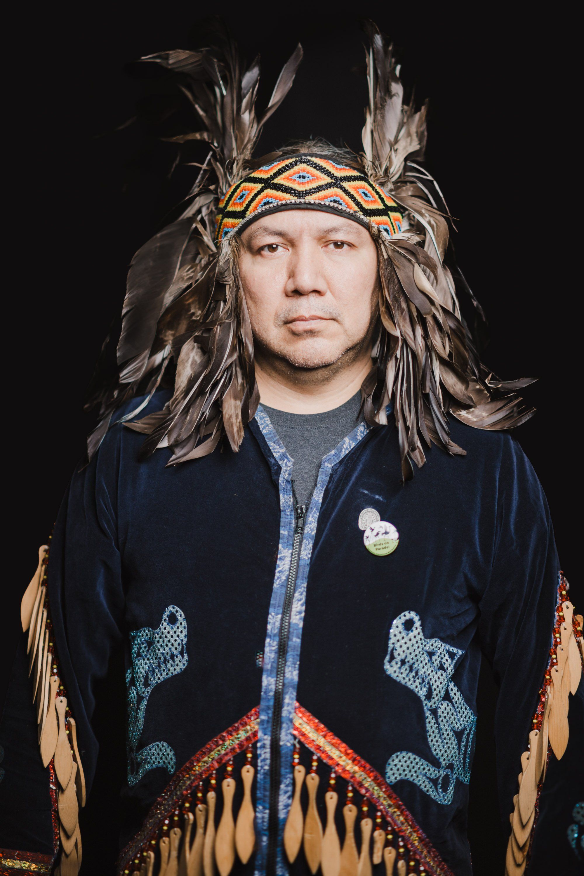 Portrait of a person wearing an elaborate feathered headdress and fringed regalia against a black background, gazing directly at camera with solemn expression