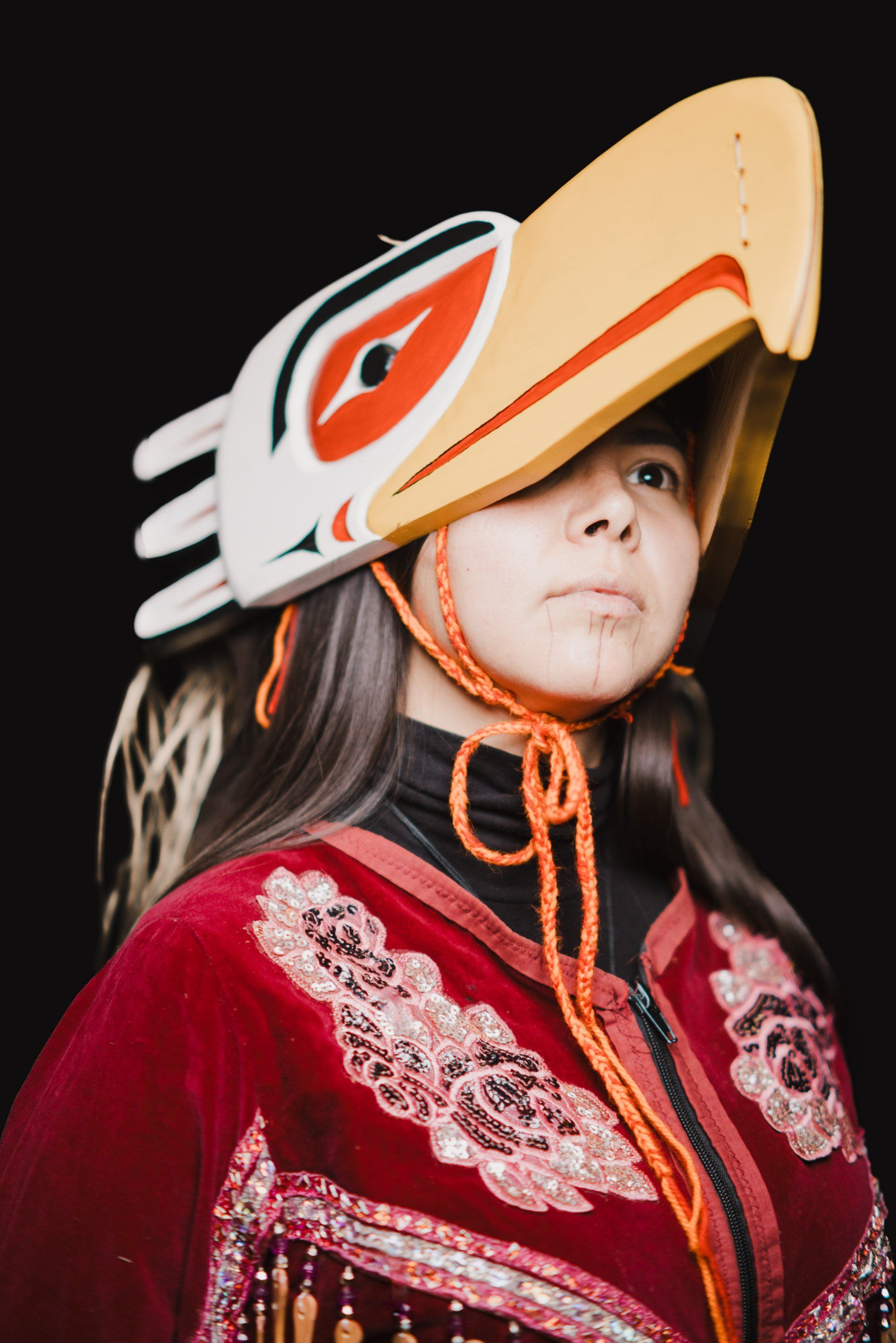 Woman wearing traditional red regalia and a carved wooden raven headdress looks upward against a black background