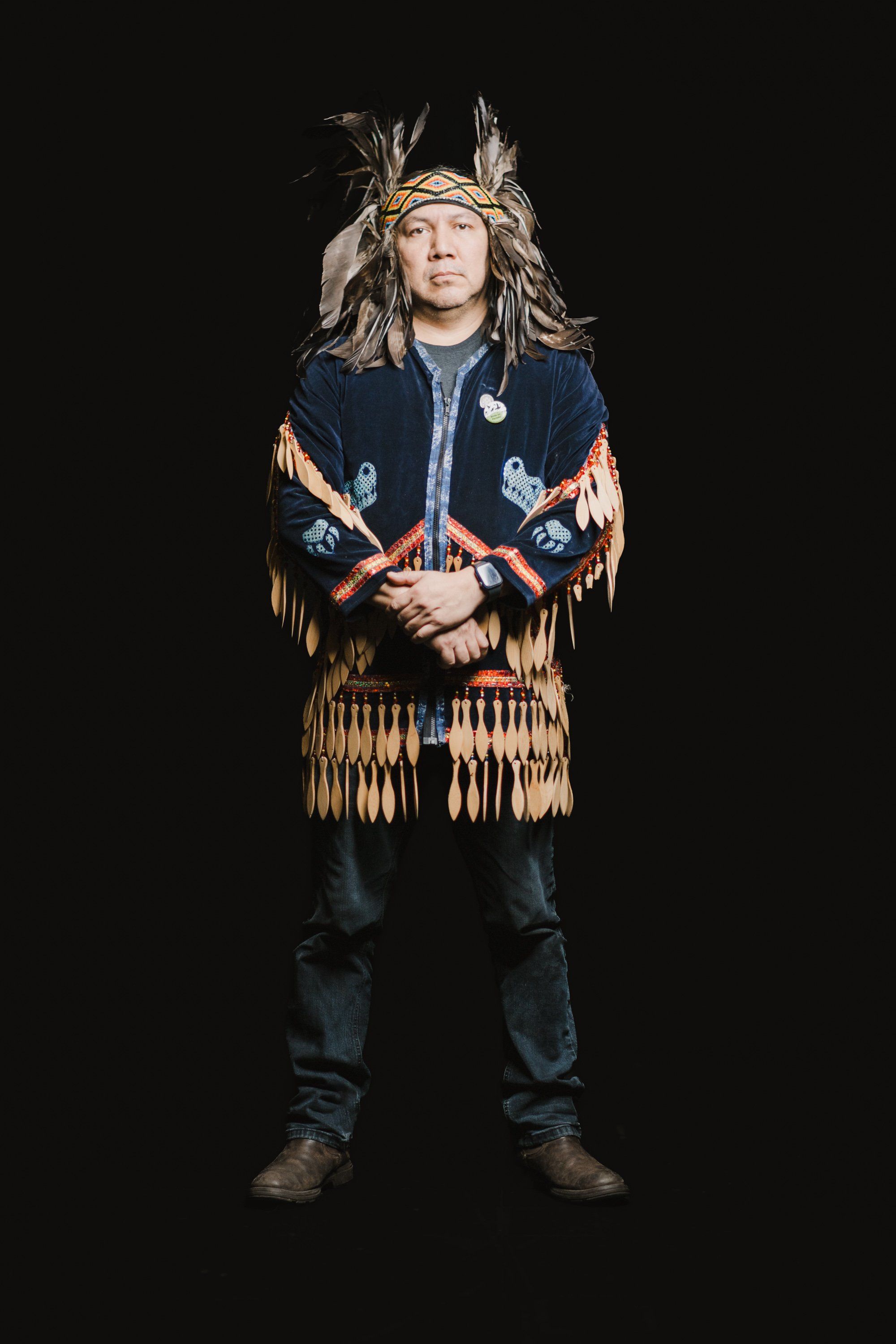 Indigenous performer in traditional Coast Salish regalia with feathered headdress and fringed leather vest stands against black background, arms crossed