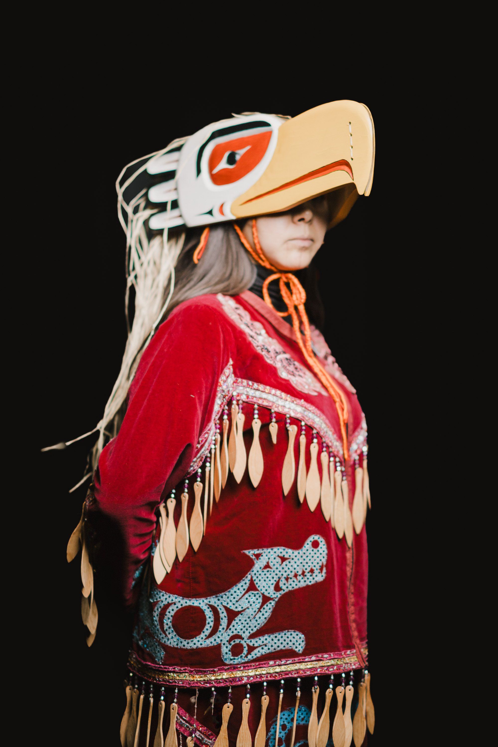 Young dancer in striking red regalia with cream eagle headdress and fringe, posed against black backdrop