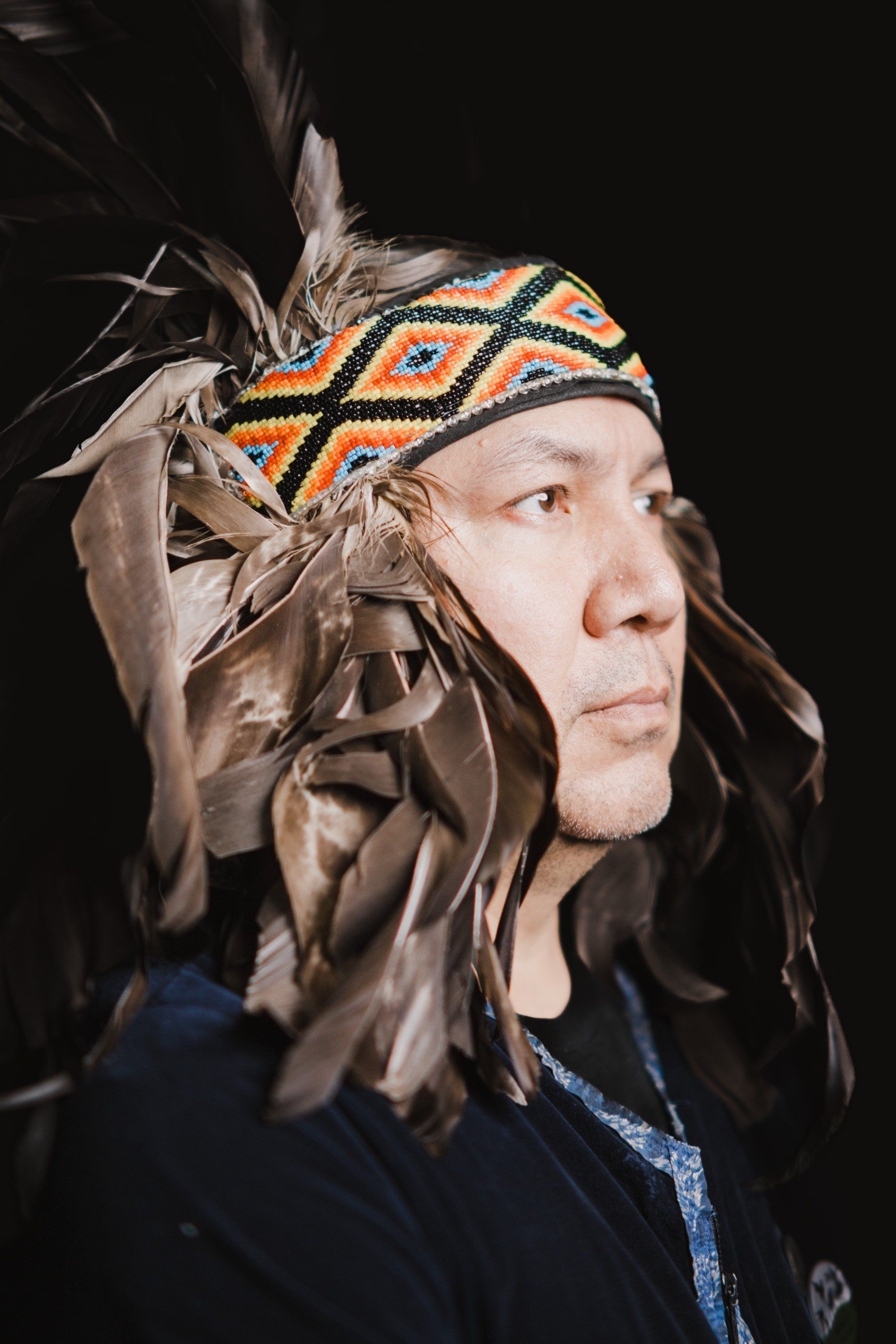 Coast Salish performer in traditional regalia with feathered headdress and beaded headband, photographed in profile against black background