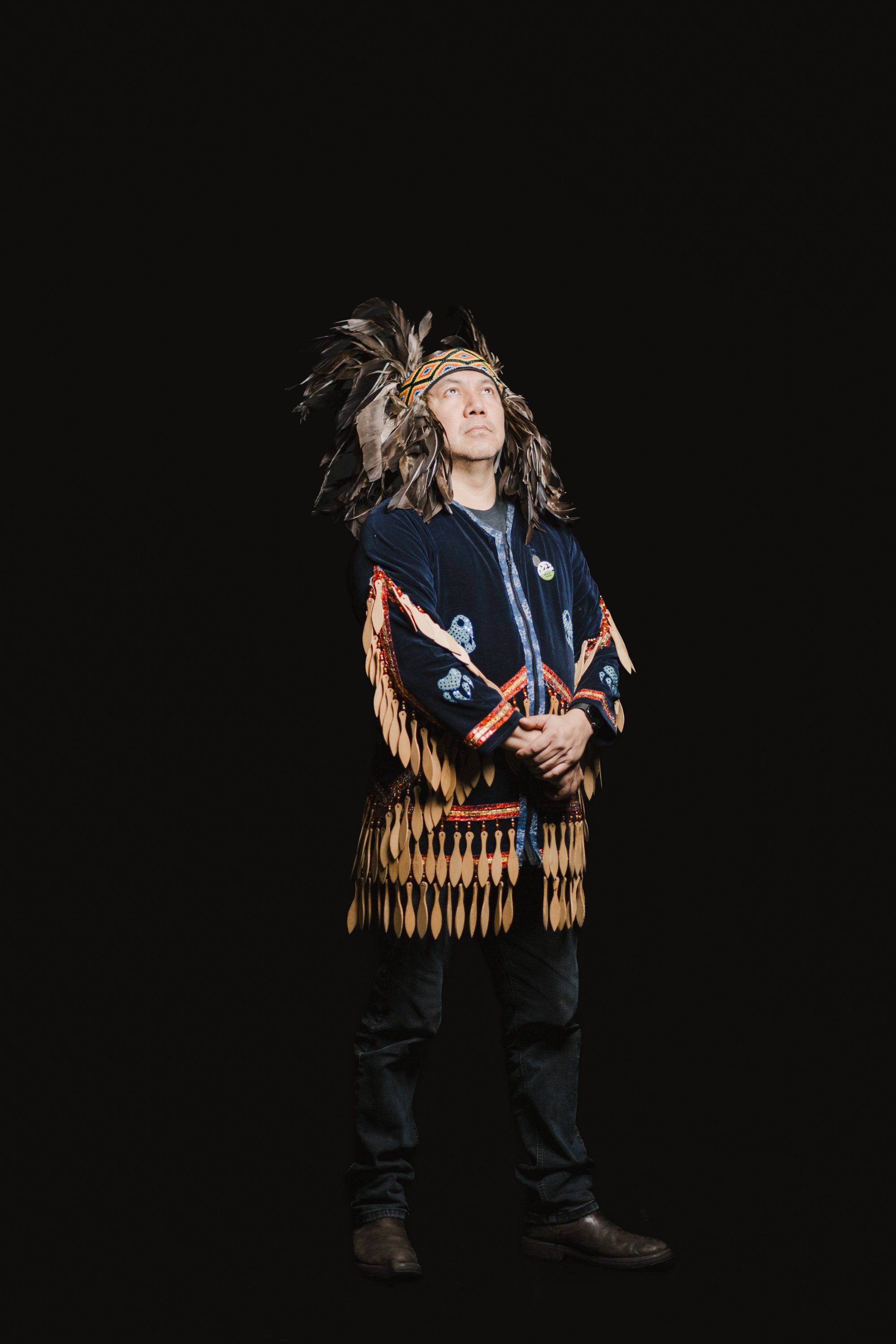 Performer in traditional Coast Salish regalia with fringed tunic and headdress gazes upward against black background