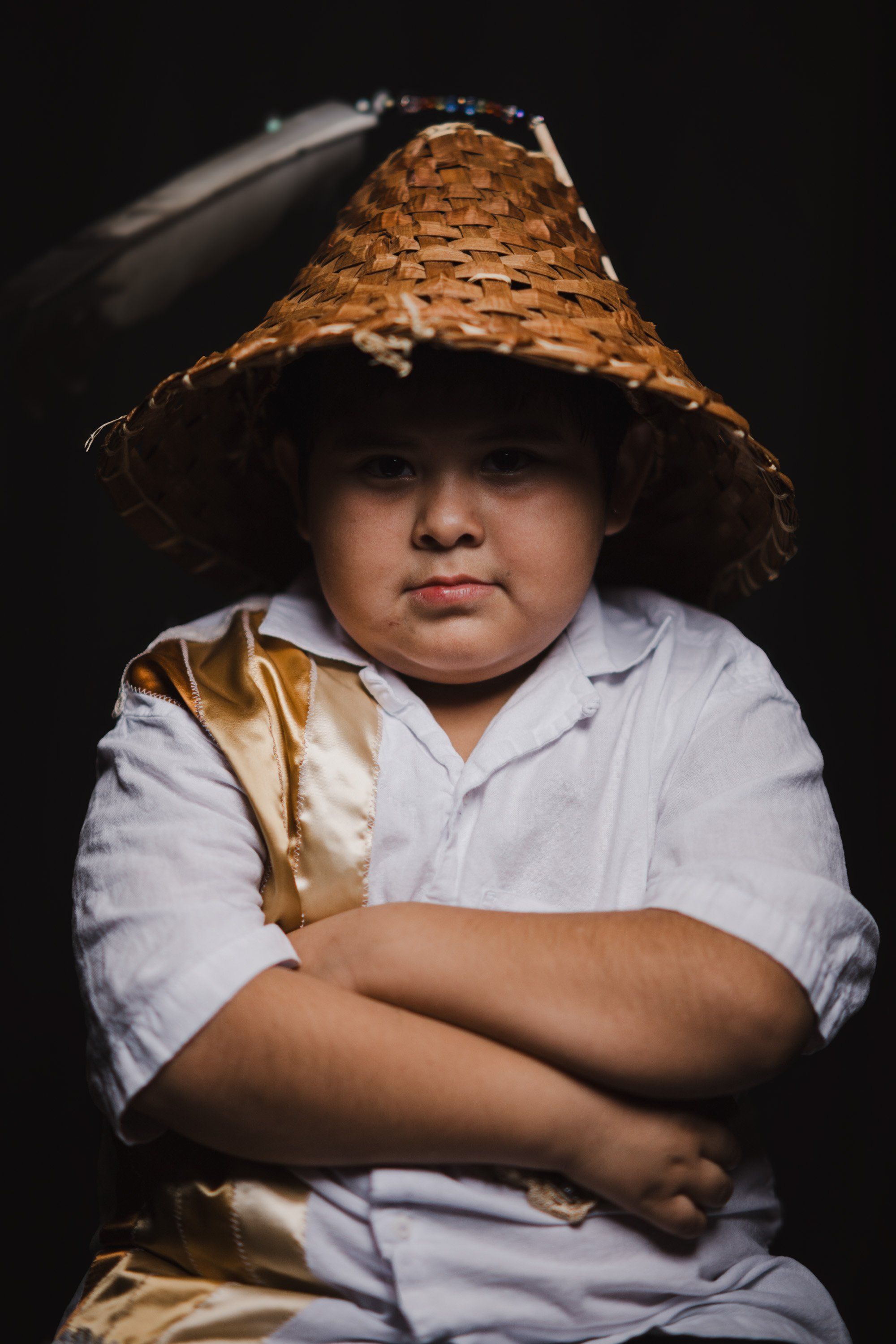Young person in white shirt and golden-brown woven hat with arms crossed, gazing directly at camera against dark background