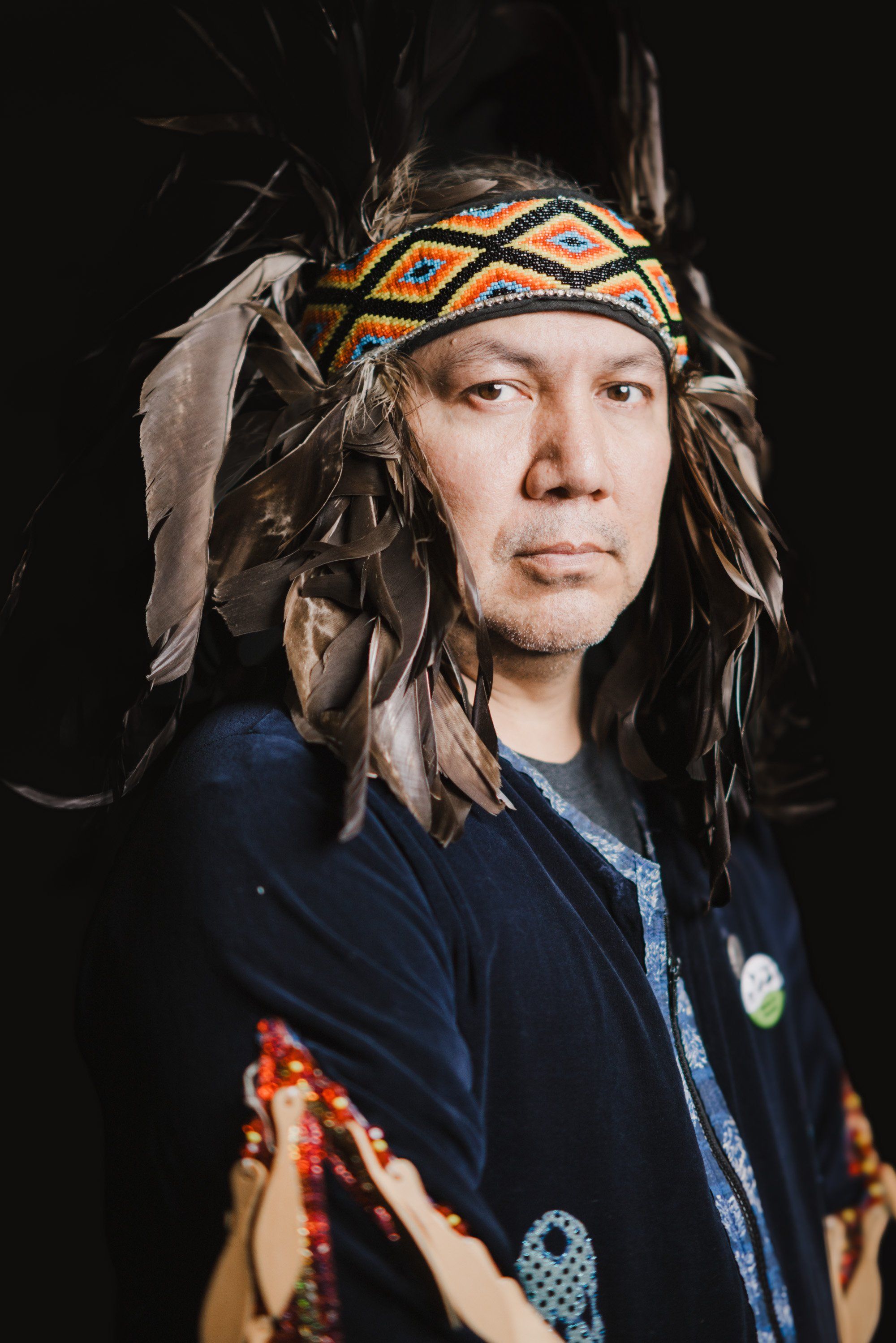 Portrait of a man wearing traditional Coast Salish regalia including a feathered headdress and decorated button blanket against a dark background