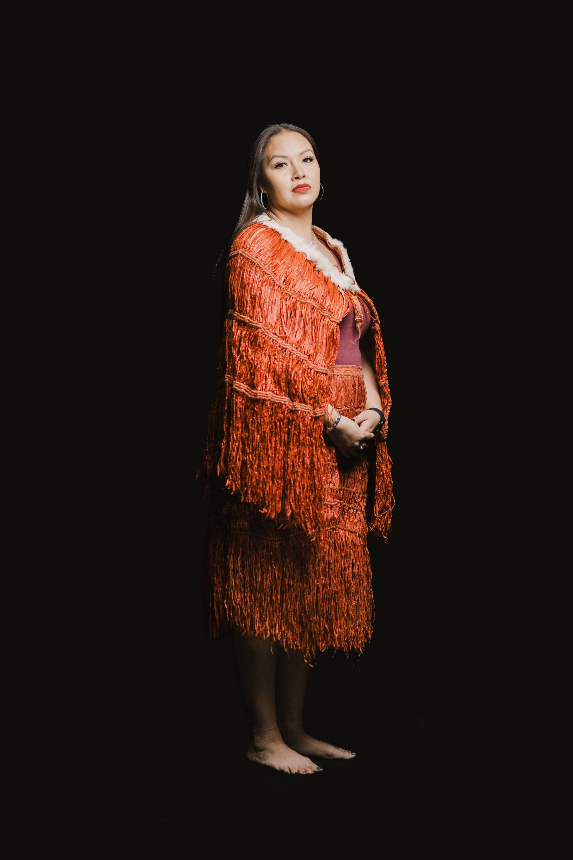 Woman in ornate orange fringed shawl stands barefoot against black background, gazing upward with serene expression
