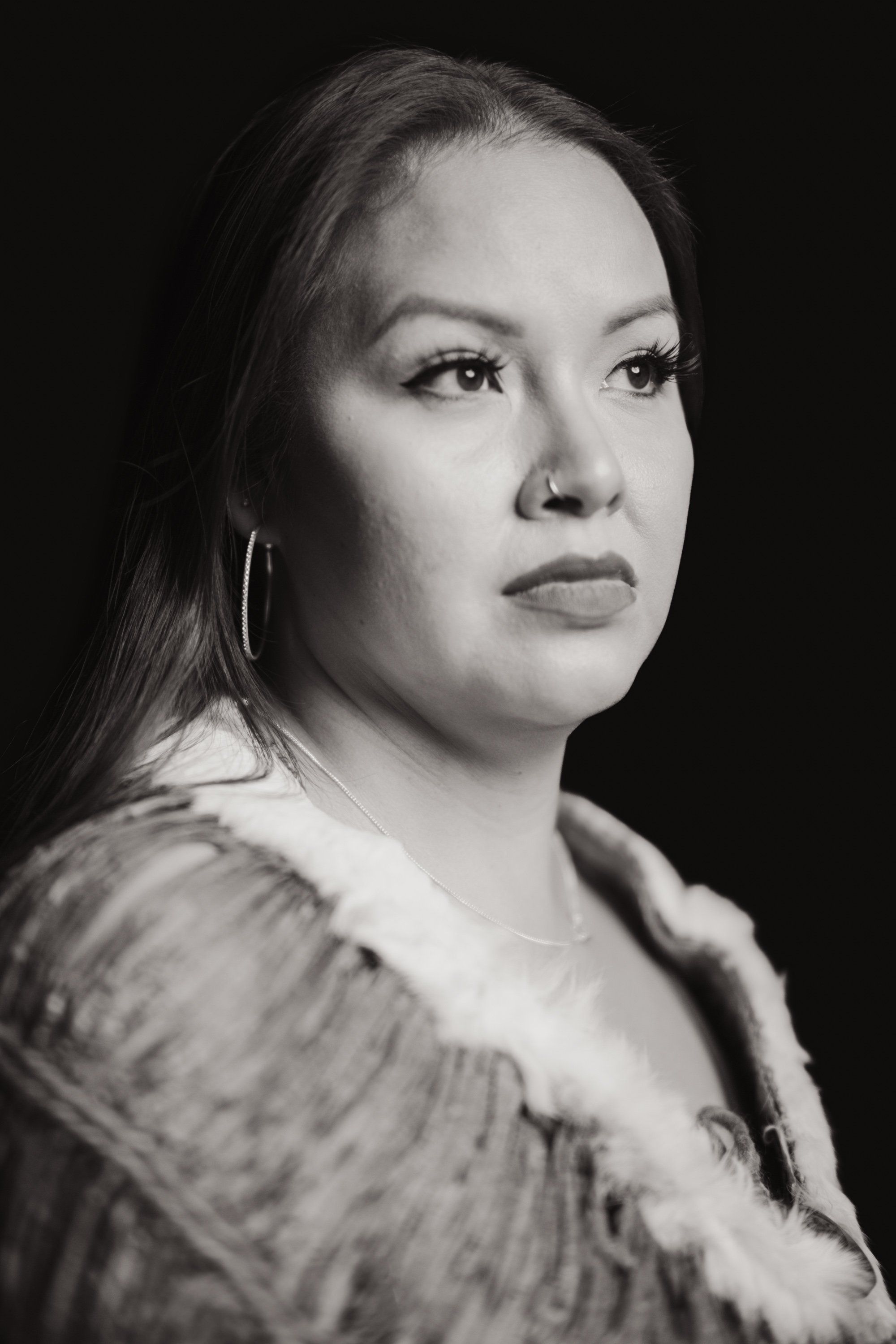 Portrait of a woman with long dark hair and hoop earrings against a black background, gazing thoughtfully to the side in dramatic studio lighting