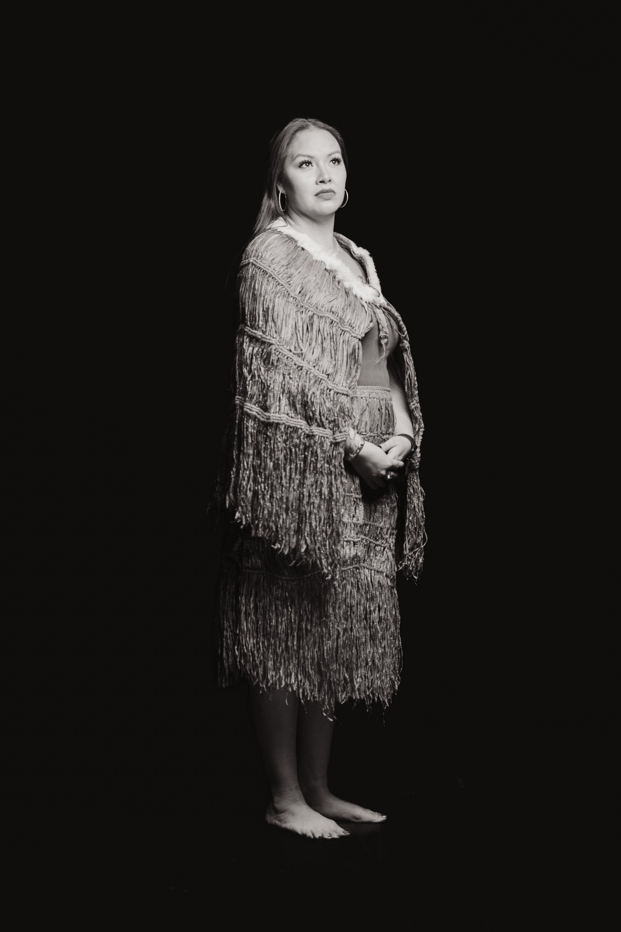 Woman in fringed shawl stands barefoot against black background, gazing upward with contemplative expression